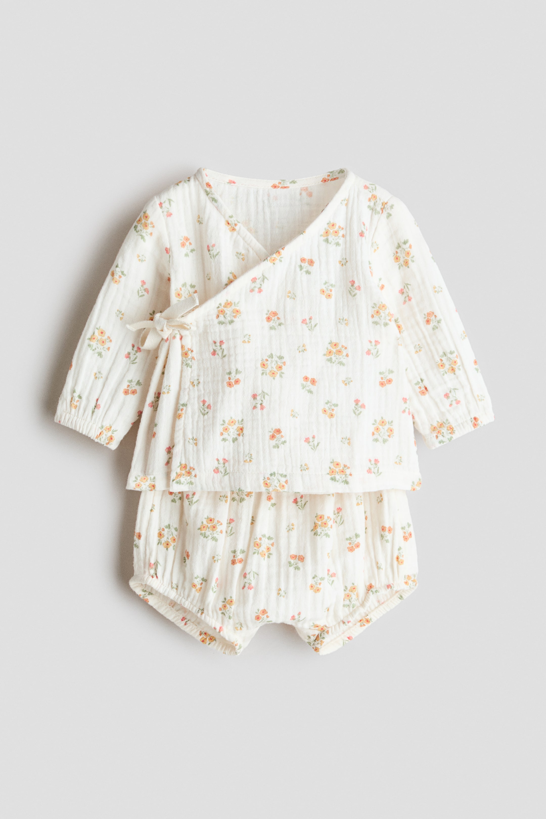 View larger image: A front view of a baby's off-white cotton muslin set, showcasing a delightful all-over floral print with petite orange and yellow blossoms. The wrap-over top presents a V-neck, long sleeves with elasticated cuffs, and side ties, while the coordinating bloomers feature an elasticated waist and leg openings.
