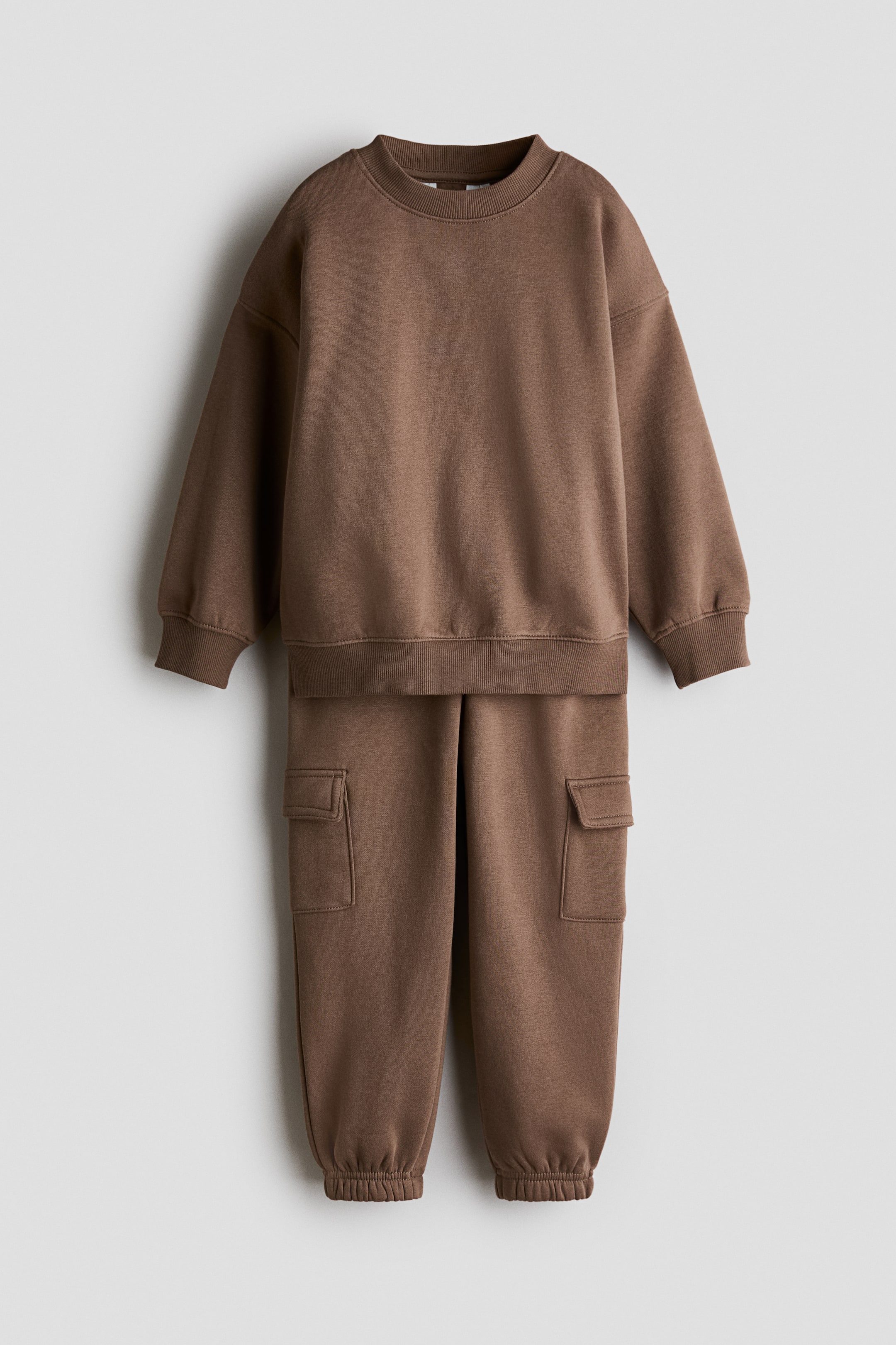 Visa större bild: Ett brunt tvådelat loungeklädesset hänger framifrån, med en sweatshirt med rund halsringning, långa ärmar, ribbade muddar och ribbad nederkant, samt matchande joggingbyxor med cargofickor på varje ben och ribbade ankelmuddar.