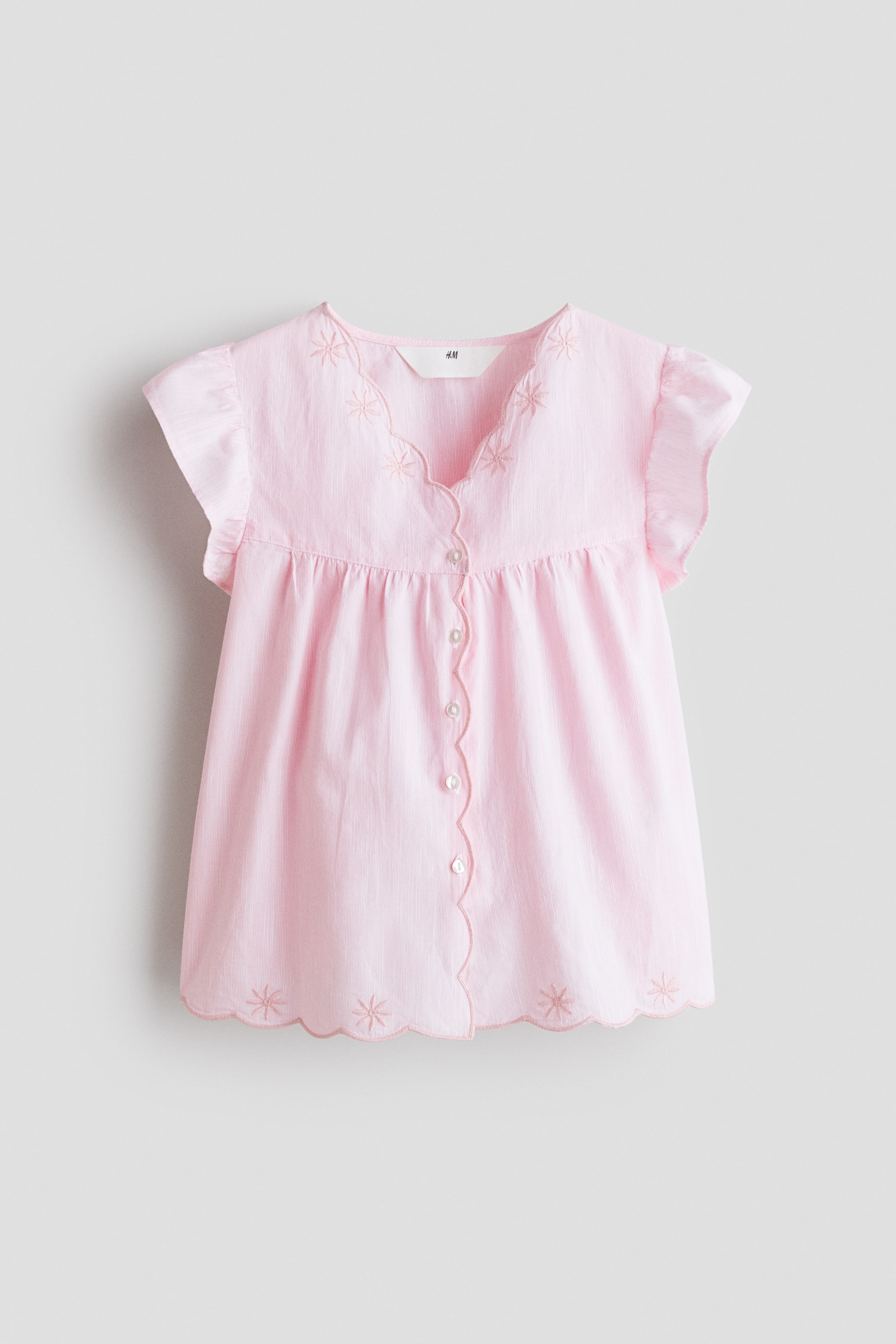 View larger image: A light pink top showcases its front, featuring a scalloped V-neckline with embroidered star motifs and short ruffled cap sleeves. The bodice is gathered, leading to a scalloped button placket with small white buttons, and a matching scalloped hem with embroidered star patterns.