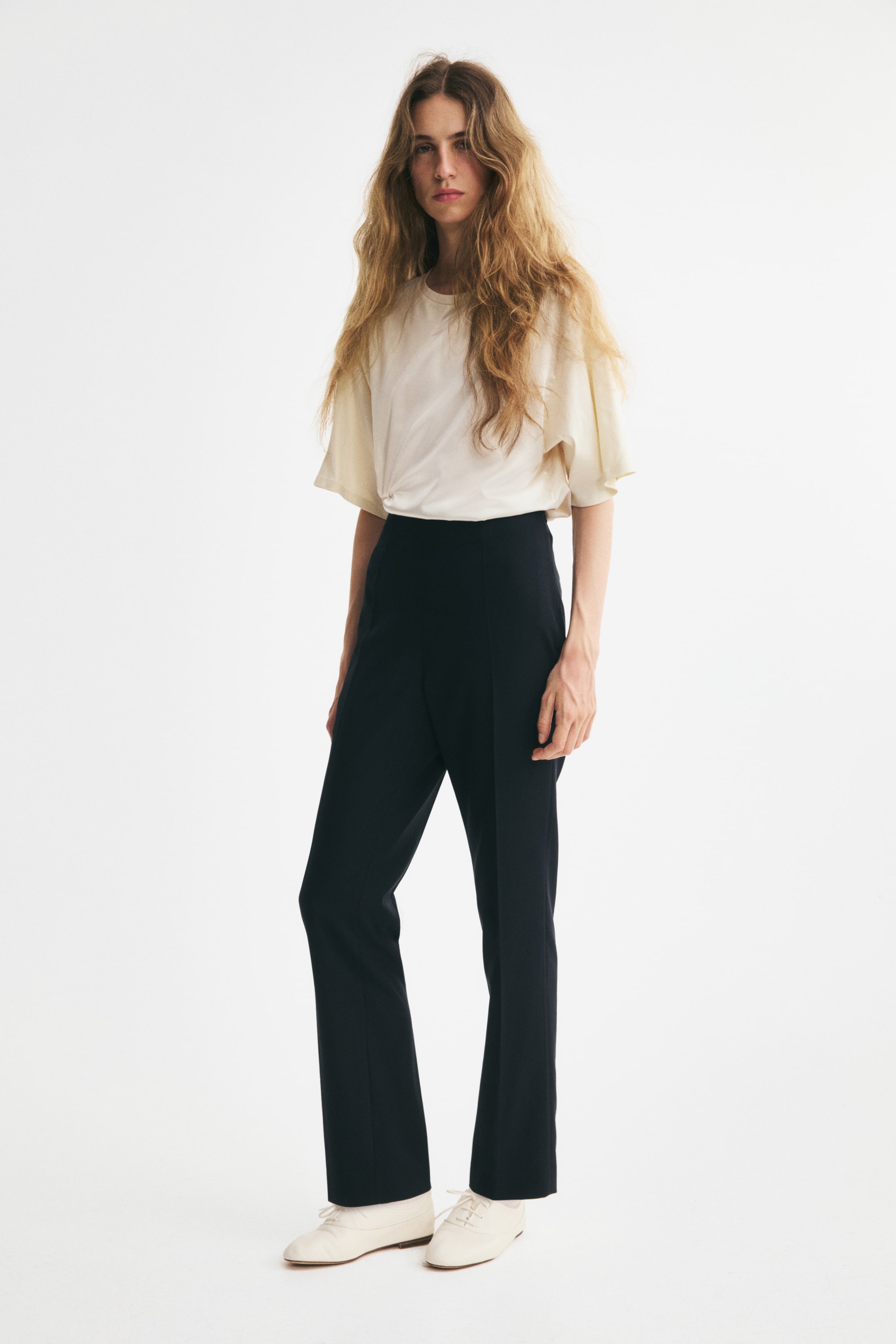 View larger image: Tailored black twill pants are worn by a model standing at a three-quarter front angle, showcasing a high-waisted fit and slim legs with visible creases running down the front. The pants are styled with a tucked-in cream-colored T-shirt to highlight the clean waistline.