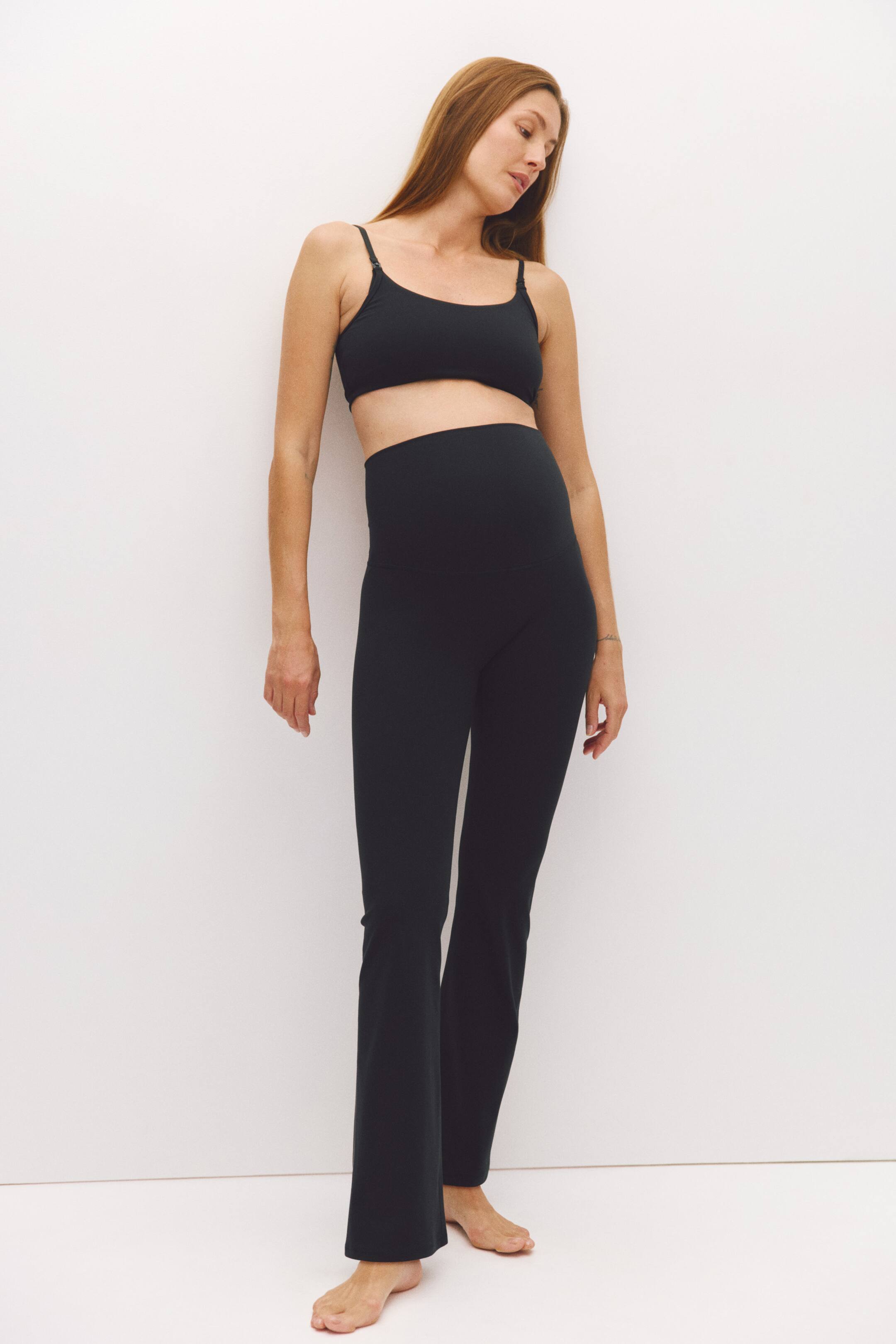 Ver imagen más grande: La modelo lleva un conjunto deportivo premamá negro, compuesto por pantalones acampanados de cintura alta y un bralette a juego con escote redondo. Los pantalones cuentan con una cinturilla de soporte por encima de la barriga que cubre el vientre embarazado de la modelo.