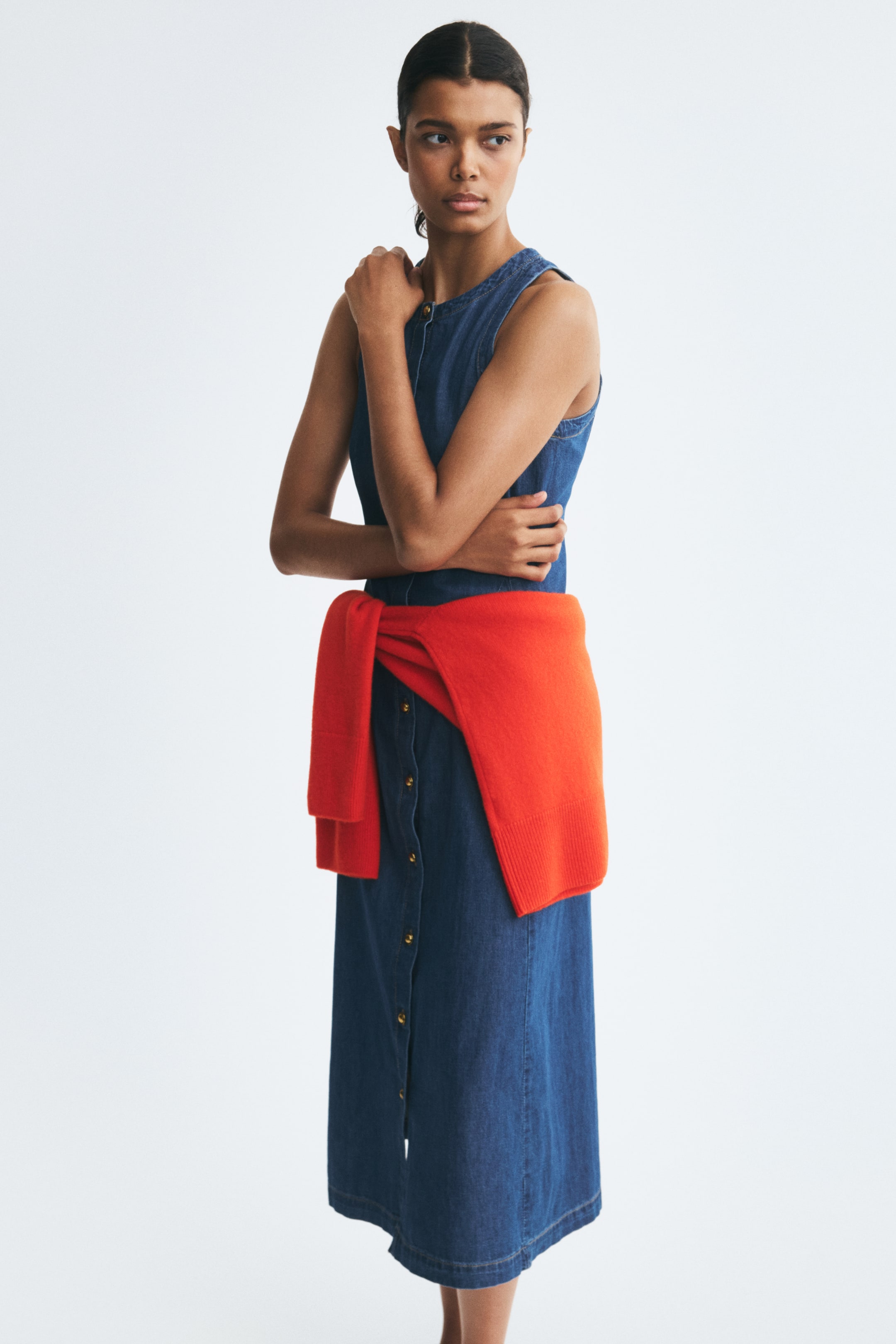 View larger image: The model is wearing a sleeveless, calf-length blue denim dress with a round neckline and a button-front closure. A bright orange-red sweater is tied around the waist.