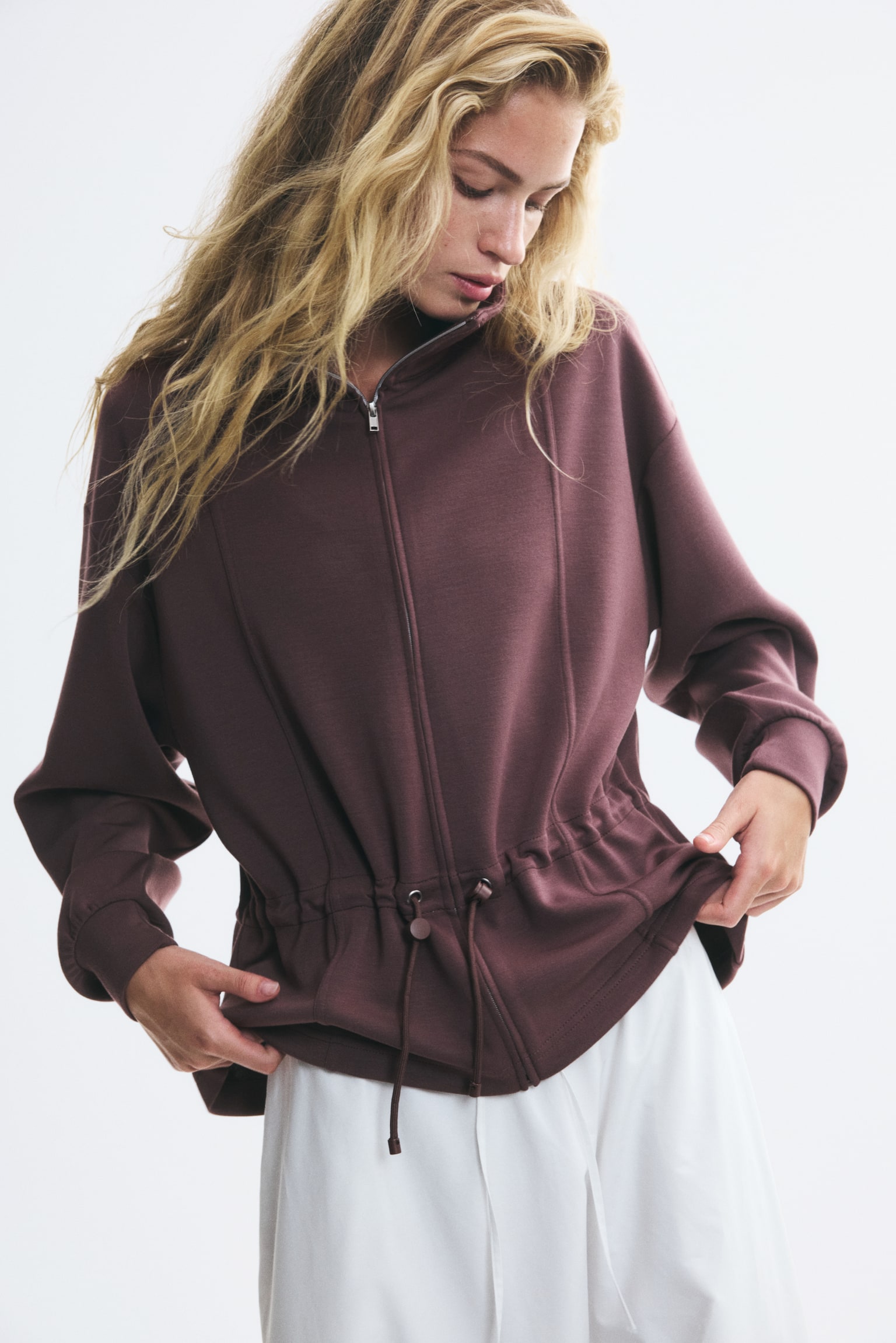 A dark brown sweat fabric sweater featuring a stand collar, a zip-up front with vertical seams, and an adjustable drawstring waist is being worn by the model. The model is holding the flared hem of the sweater, which is paired with white trousers. - 1