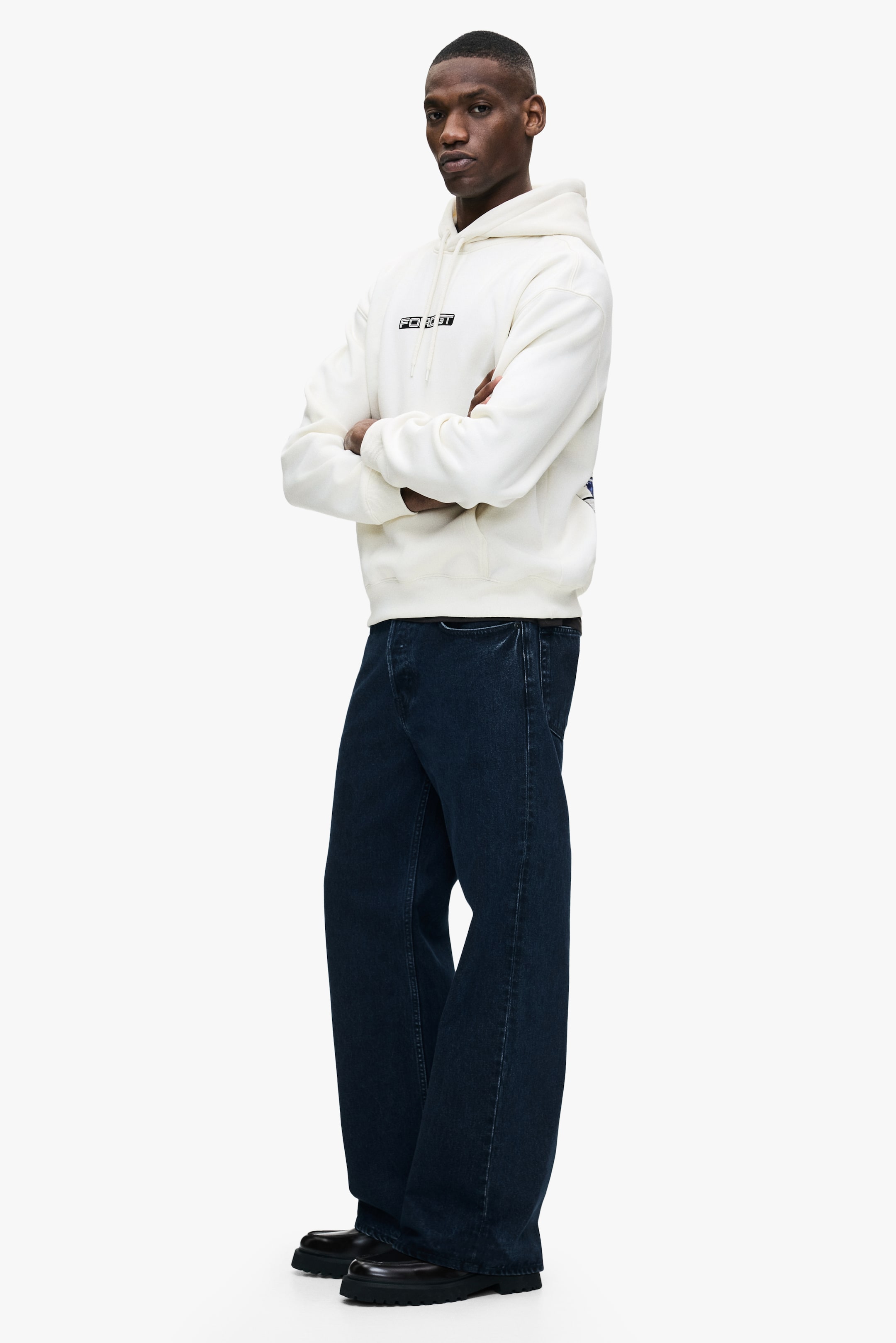 Größeres Bild anzeigen: Ein cremefarbener Hoodie mit schwarzem Grafiktext auf der Brust, einer Kapuze und einer Fronttasche wird vom Model getragen. Die Arme des Models sind vorne verschränkt und für das Styling werden dunkel gewaschene Jeans im weiten Bein getragen.