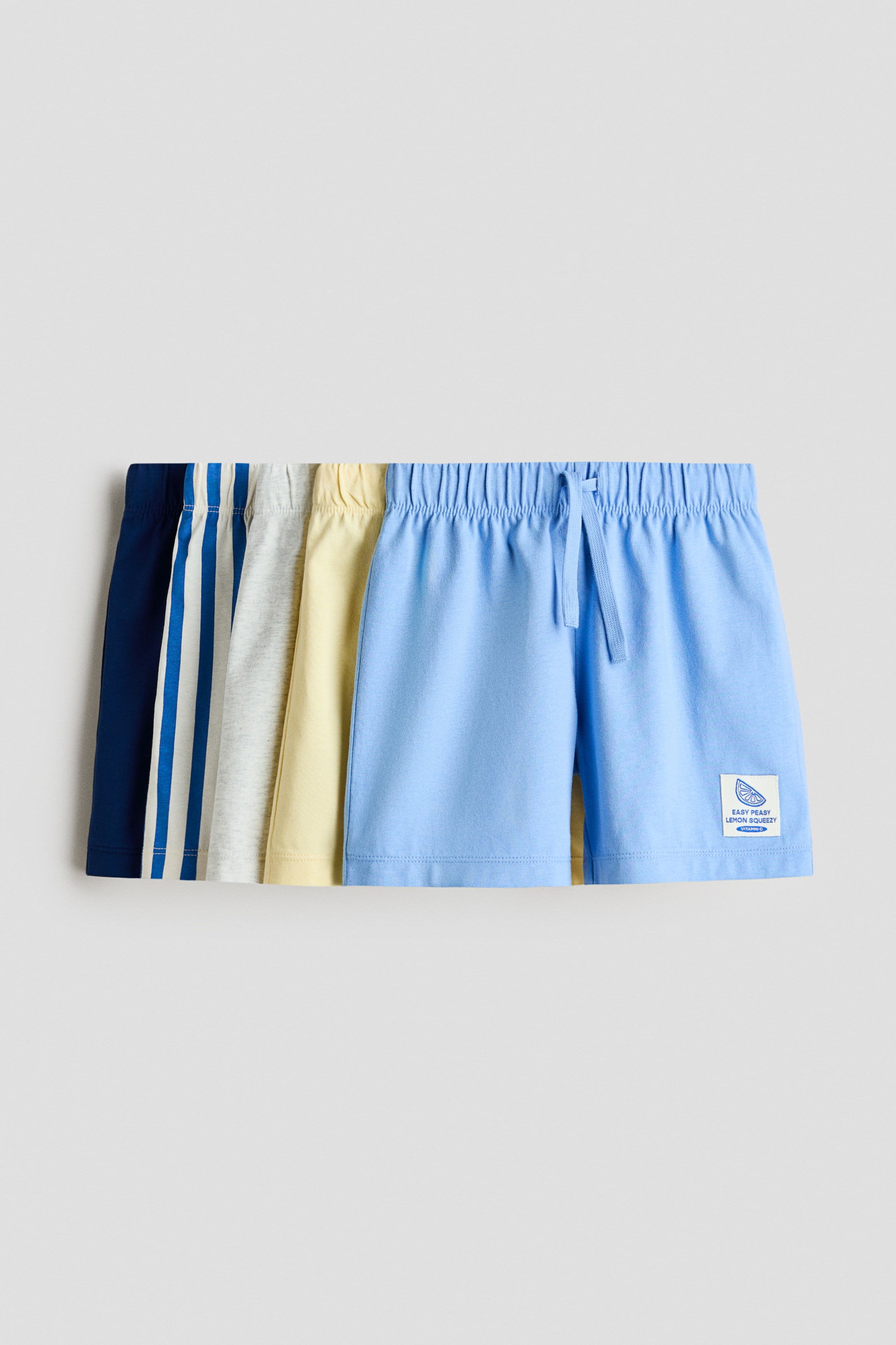 Vezi imaginea mai mare: Un set de pantaloni scurți până la genunchi din jerseu de bumbac moale este afișat, având o talie ușor de tras pe corp, cu elastic acoperit și un șnur vizibil. Pantalonii scurți sunt disponibili în mai multe culori și modele, inclusiv albastru închis, dungi albastre și albe, gri deschis melanj, galben deschis și albastru deschis, iar perechea albastru deschis din față prezintă o etichetă mică albă cu un grafic de lămâie.