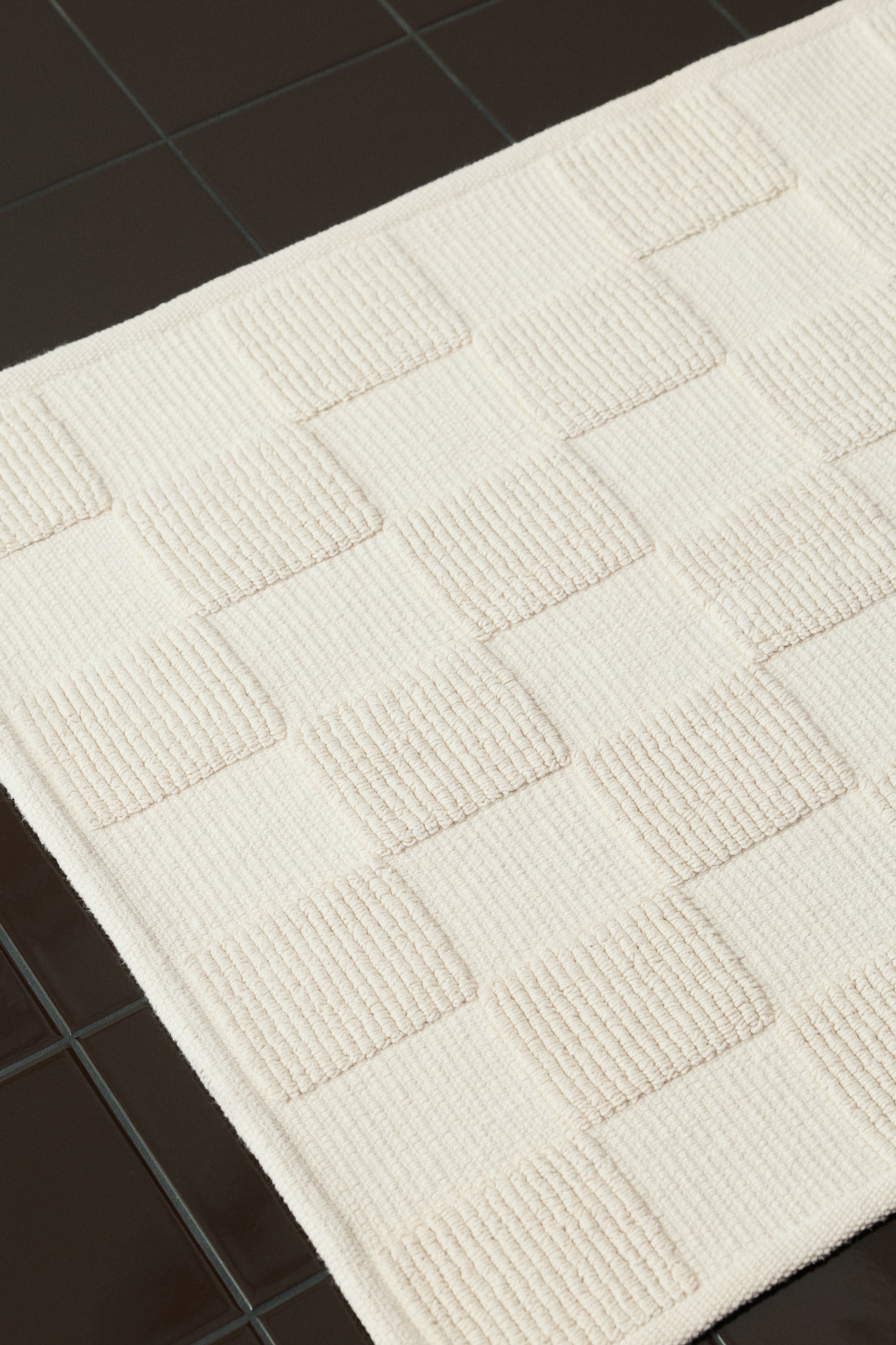 View larger image: A cream-colored bath mat is featuring a raised, geometric pattern. The design alternates horizontally ribbed, raised rectangular sections with flatter, textured squares.