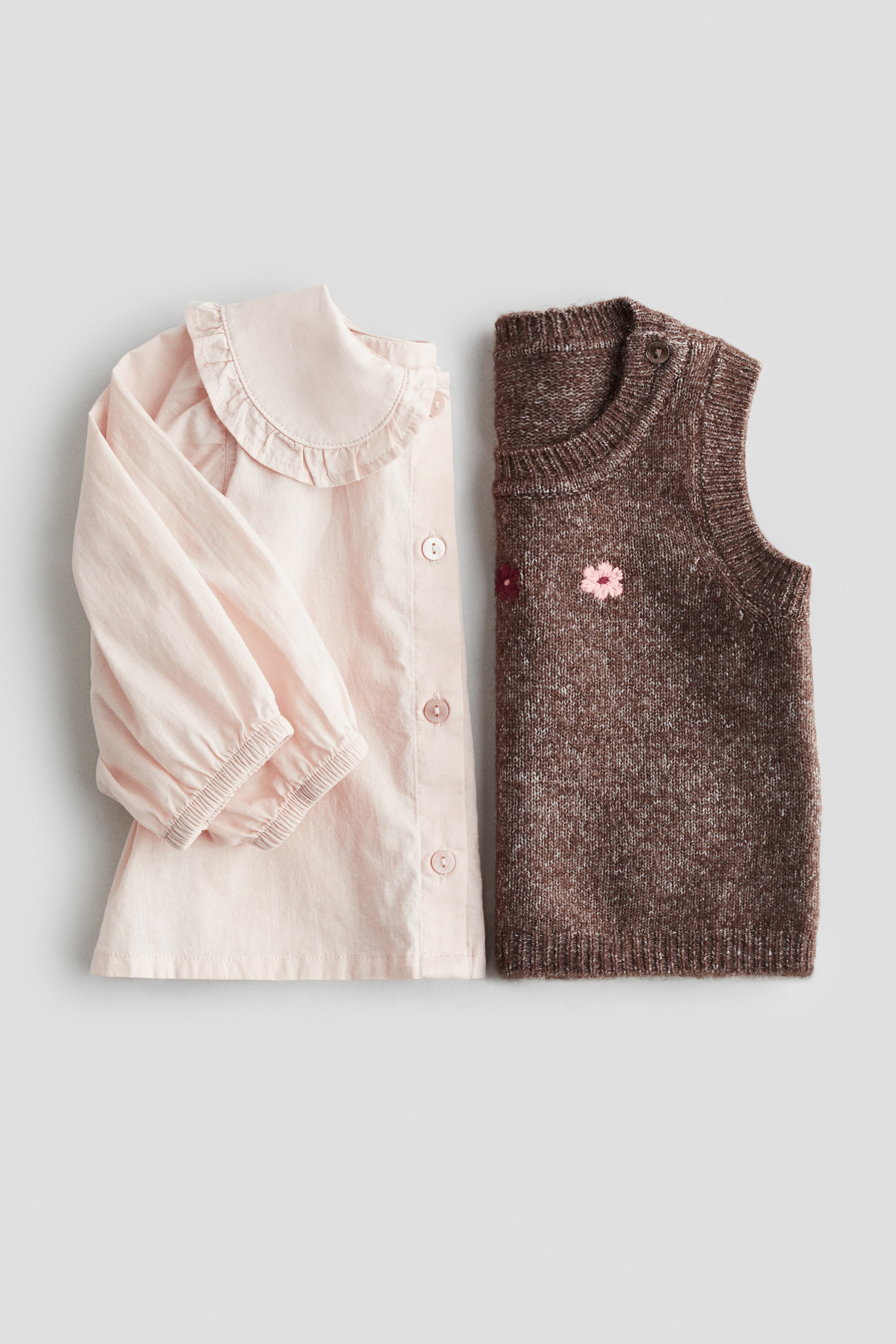 Ver imagem maior: Uma camisa de manga comprida rosa claro está deitada plana, apresentando uma gola Peter Pan com folhos, punhos franzidos e uma carcela de botões à frente com botões de cor clara visíveis. Ao lado, um colete sem mangas de malha castanha mesclada exibe uma gola redonda canelada, cavas caneladas e uma bainha canelada, com duas pequenas flores bordadas no peito e fechos de botão no ombro.