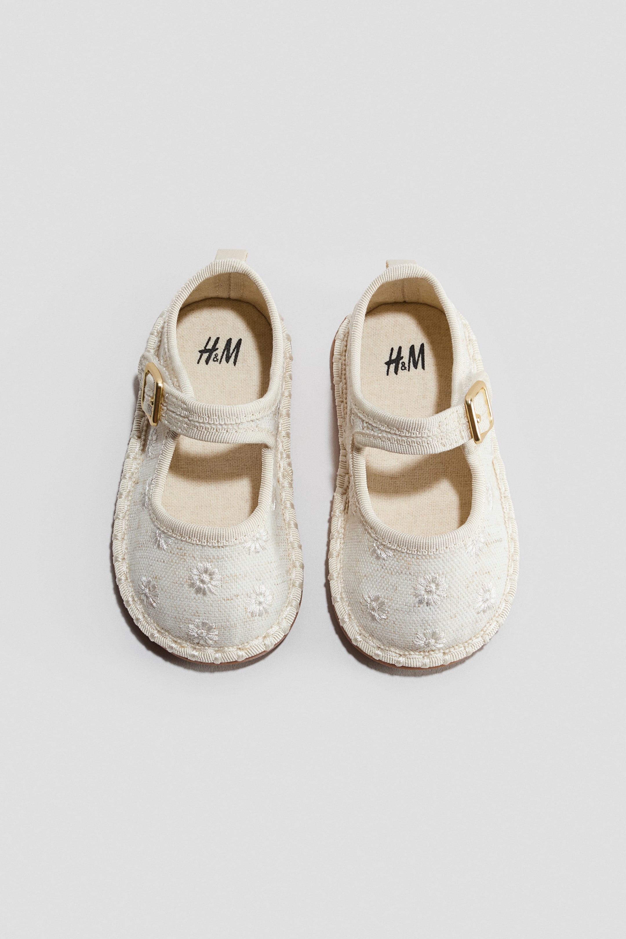 View larger image: A pair of cream baby Mary Jane shoes features intricate embroidered floral patterns across the upper. Each shoe is designed with an instep strap, fastening with a small gold-toned buckle, and showcasing braided detailing along the sole edge.