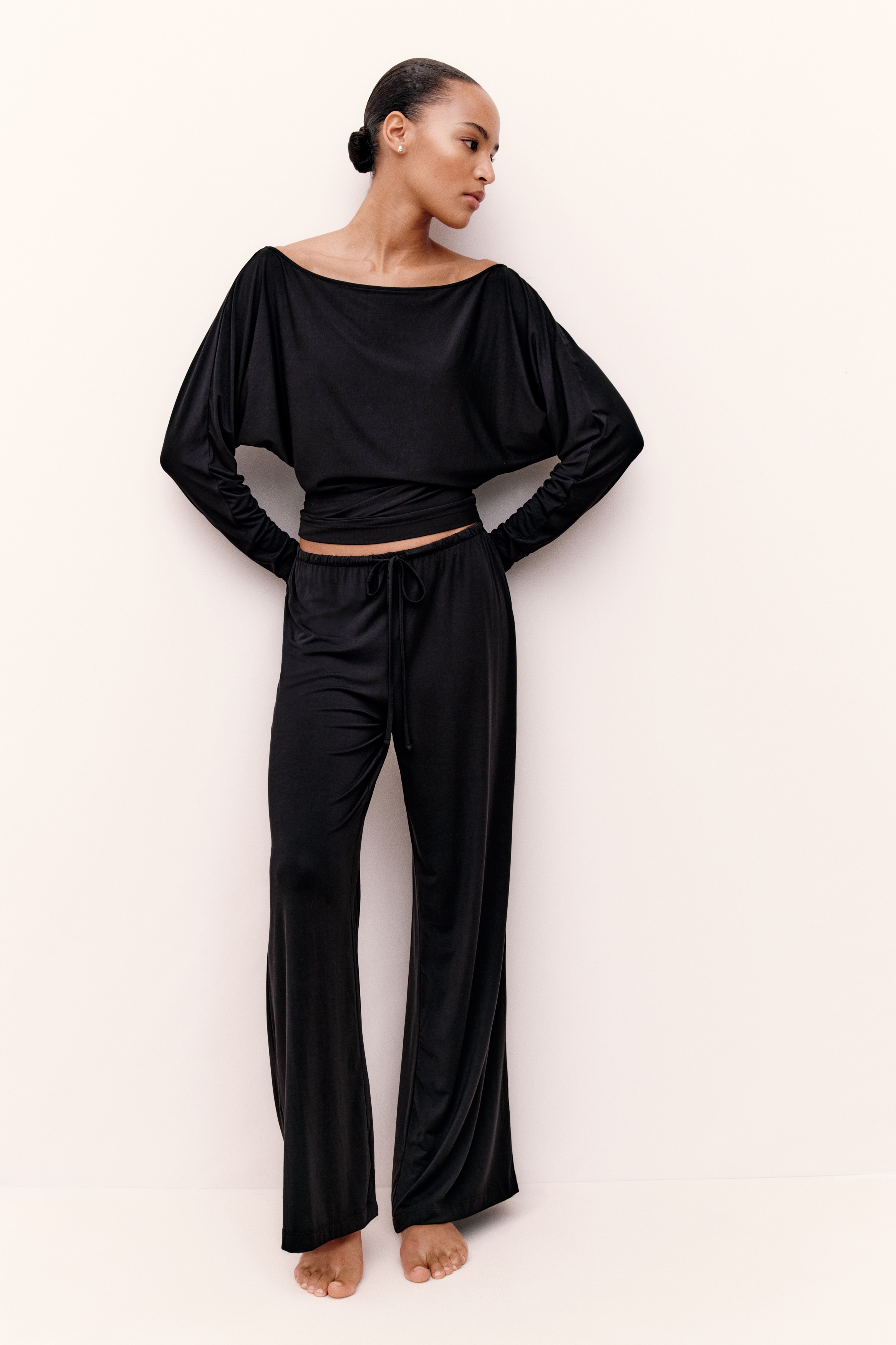 Un completo nero due pezzi Ã¨ mostrato frontalmente, composto da un top a maniche lunghe con scollo a barca drappeggiato e pantaloni coordinati a gamba larga. Il top presenta una silhouette morbida con drappeggi fluidi sul busto, mentre i pantaloni cadono con una vestibilitÃ  ampia e dritta.