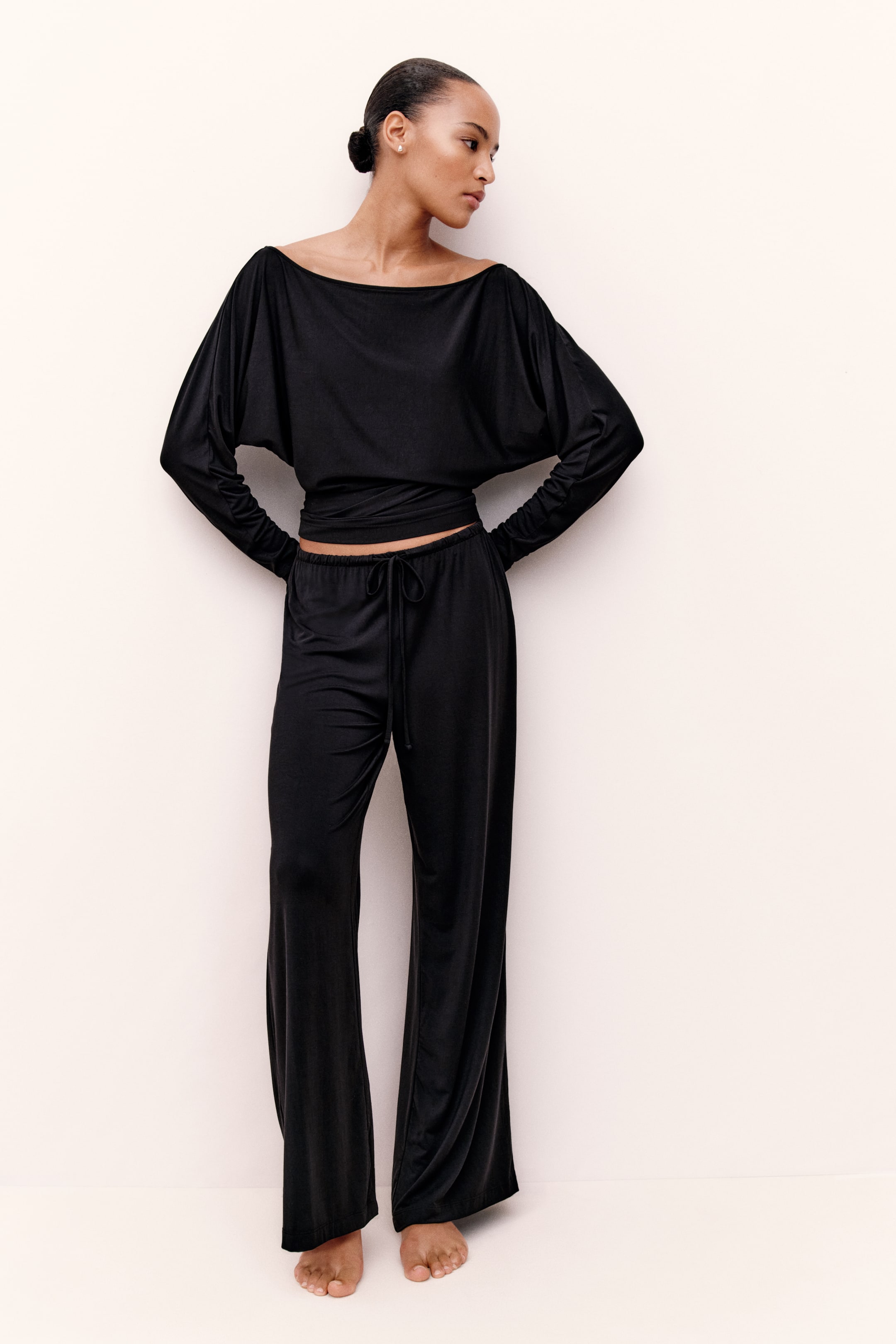 View larger image: A black pyjama set is being worn by the model, featuring a top with a boat neckline and draped dolman sleeves. The matching bottoms have a wide-leg fit and an elasticated waistband with a front drawstring tie.