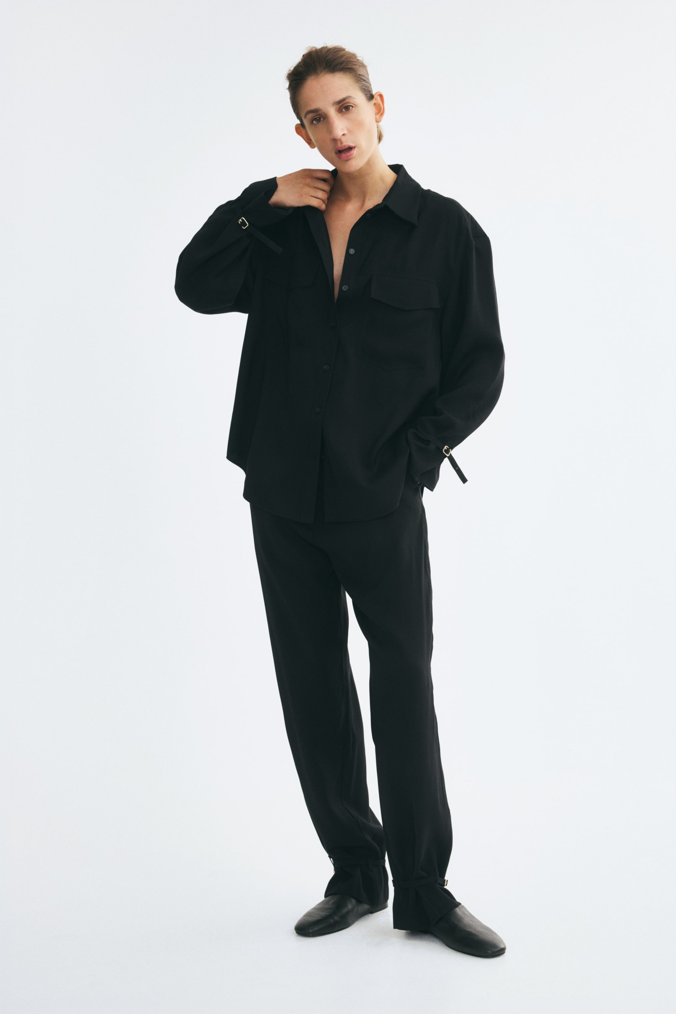 View larger image: The model is wearing black pants in a viscose-blend weave, featuring pressed creases down the front and narrow straps with metal buckles at the hems for an adjustable fit. The pants are styled with a matching black long-sleeved shirt and black flat shoes.