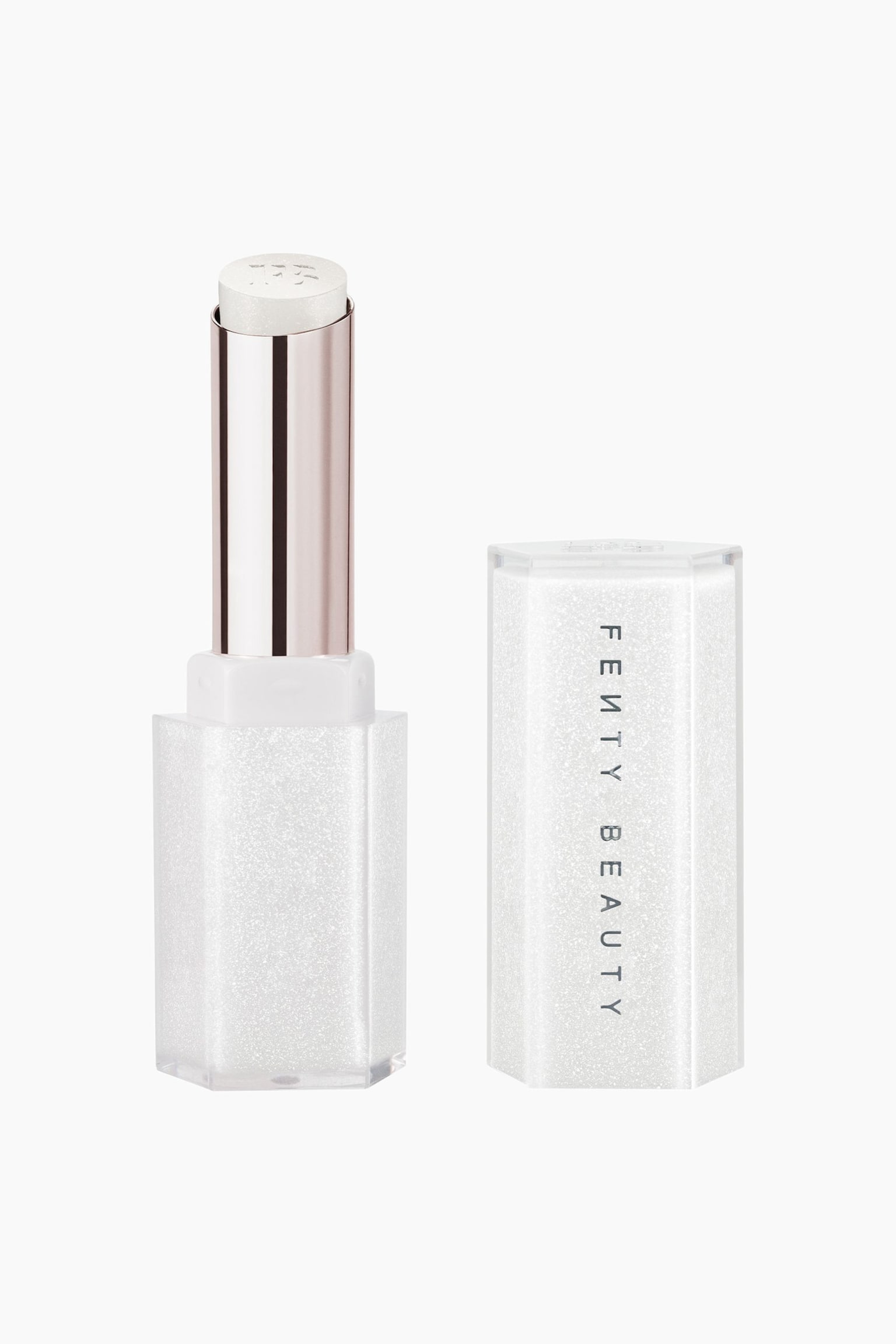 Gloss Bomb Stix High-shine Shimmer Gloss Stick stÃ¥r upprÃ¤tt med korken av och placerad till hÃ¶ger. Det Ã¶ppna glossstiftet visar upp en skimrande vit formula med glitter i en metallisk tub med en glittrig sexkantig vit bas, medan den matchande sexkantiga korken visar varumÃ¤rket "FENTY BEAUTY" vertikalt.