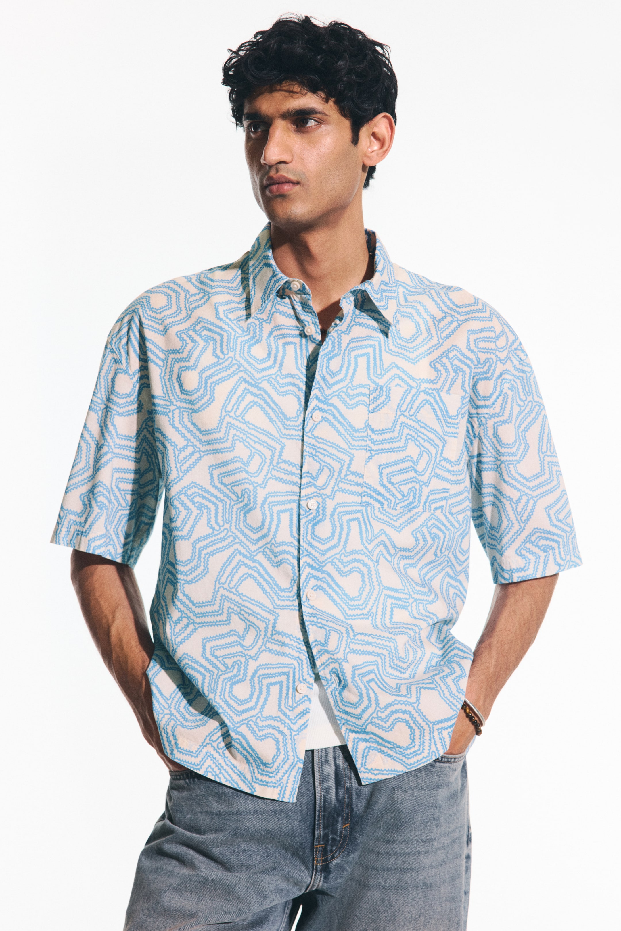 View larger image: The shirt, with short sleeves and a button-front design, is displayed from the front with a slight right angle, highlighting its relaxed fit. It boasts a light cream base adorned with a repeating light blue abstract wavy line pattern, a pointed collar, and a single left chest patch pocket. A white undershirt is visible underneath, adding to the overall look.