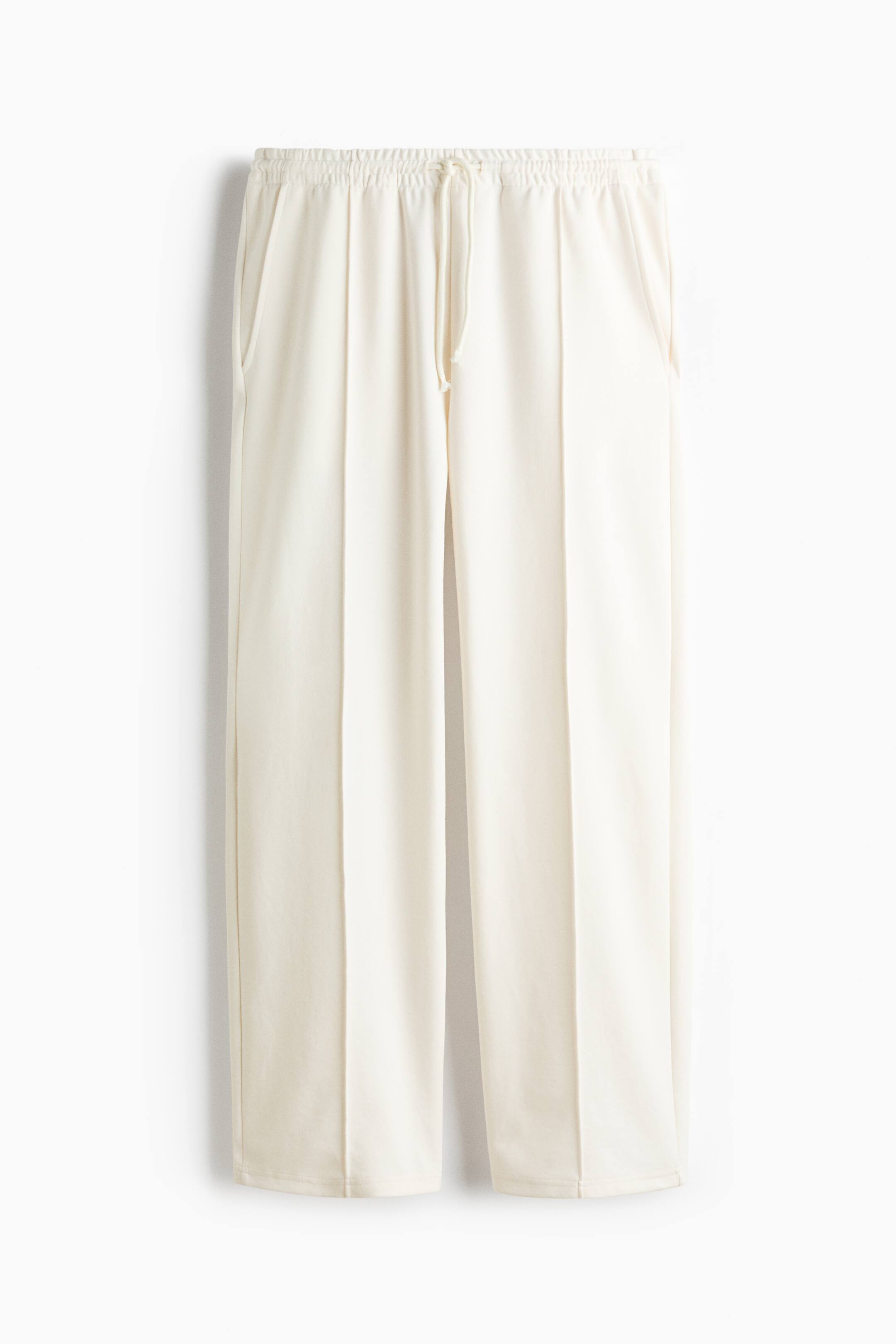 View larger image: The cream-coloured trousers feature an elasticated waistband with a visible drawstring tie at the front. They also display side seam pockets and prominent vertical creases running down the front of the straight legs.