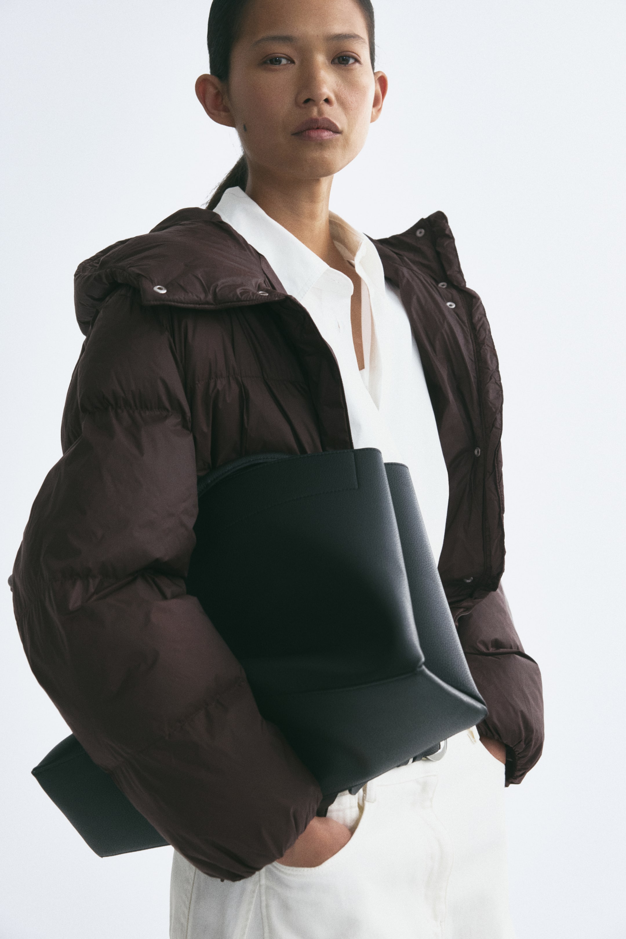 View larger image: A dark brown hooded puffer jacket with a quilted design is worn open by the model over a white button-down shirt and white trousers. The model is carrying a large black leather tote bag tucked under their arm.