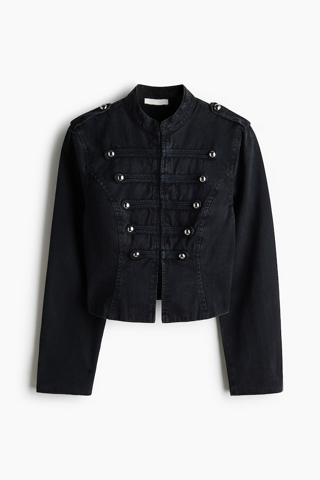 A black denim military-style jacket is facing front, featuring a band collar and shoulder epaulets with silver dome buttons. The jacket is showcasing horizontal braided frogging details across the chest accented with matching silver dome buttons and is ending in a cropped, open-front hem. - 1