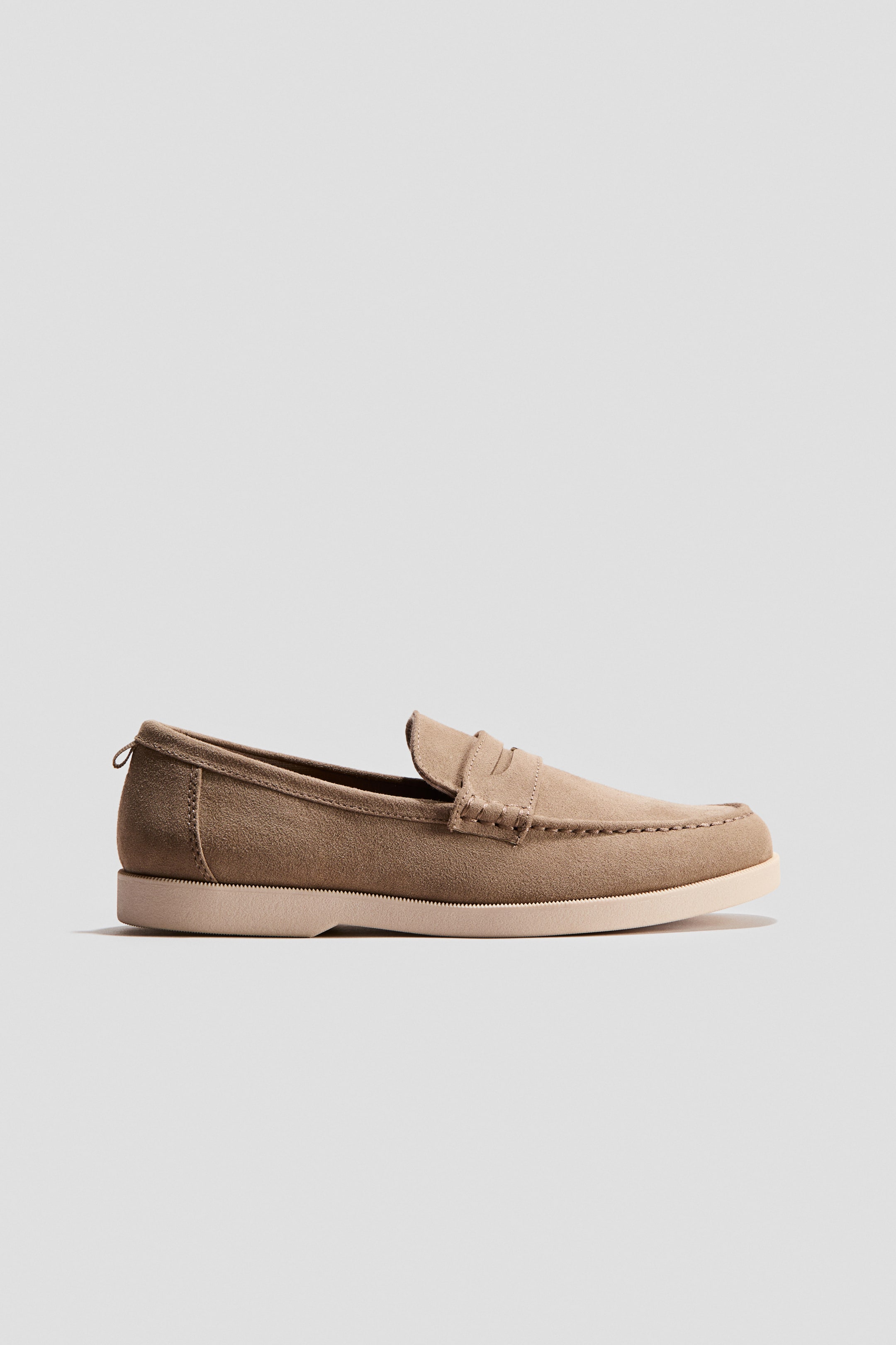 Se større billede: En tanfarvet ruskinds penny loafer vender mod højre, vinklet for at vise sin side og front. Den har mokkasinsyninger omkring tåen, en flad, lys beige sål og en lille trækstrop ved hælen.
