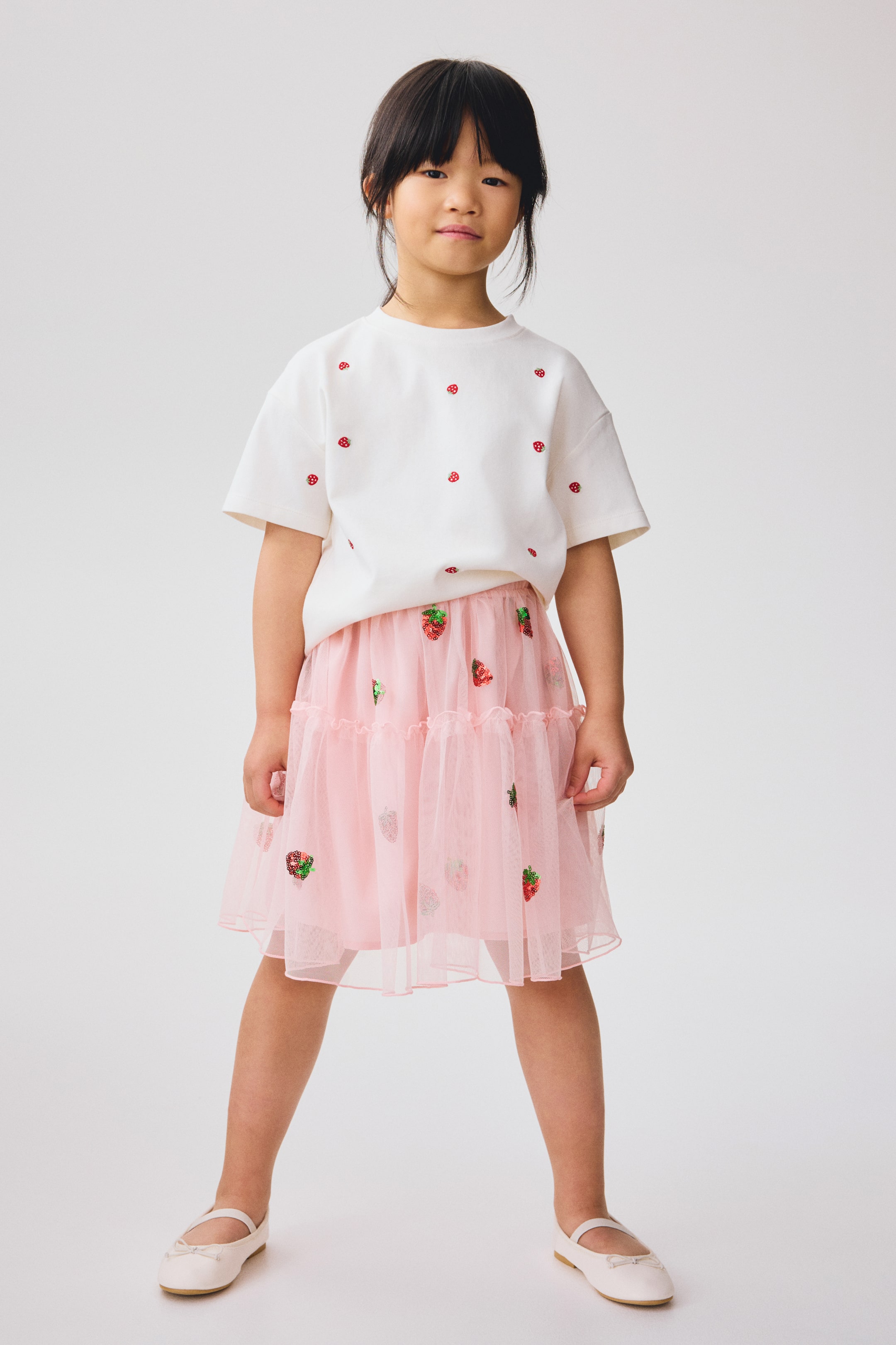 Ingrandisci l'immagine: Un top bianco a maniche corte, caratterizzato da piccole fragole rosse ricamate, è indossato sopra una gonna rosa in tulle a strati. La gonna è decorata con fragole di paillettes, e l'intero outfit è rivolto in avanti.