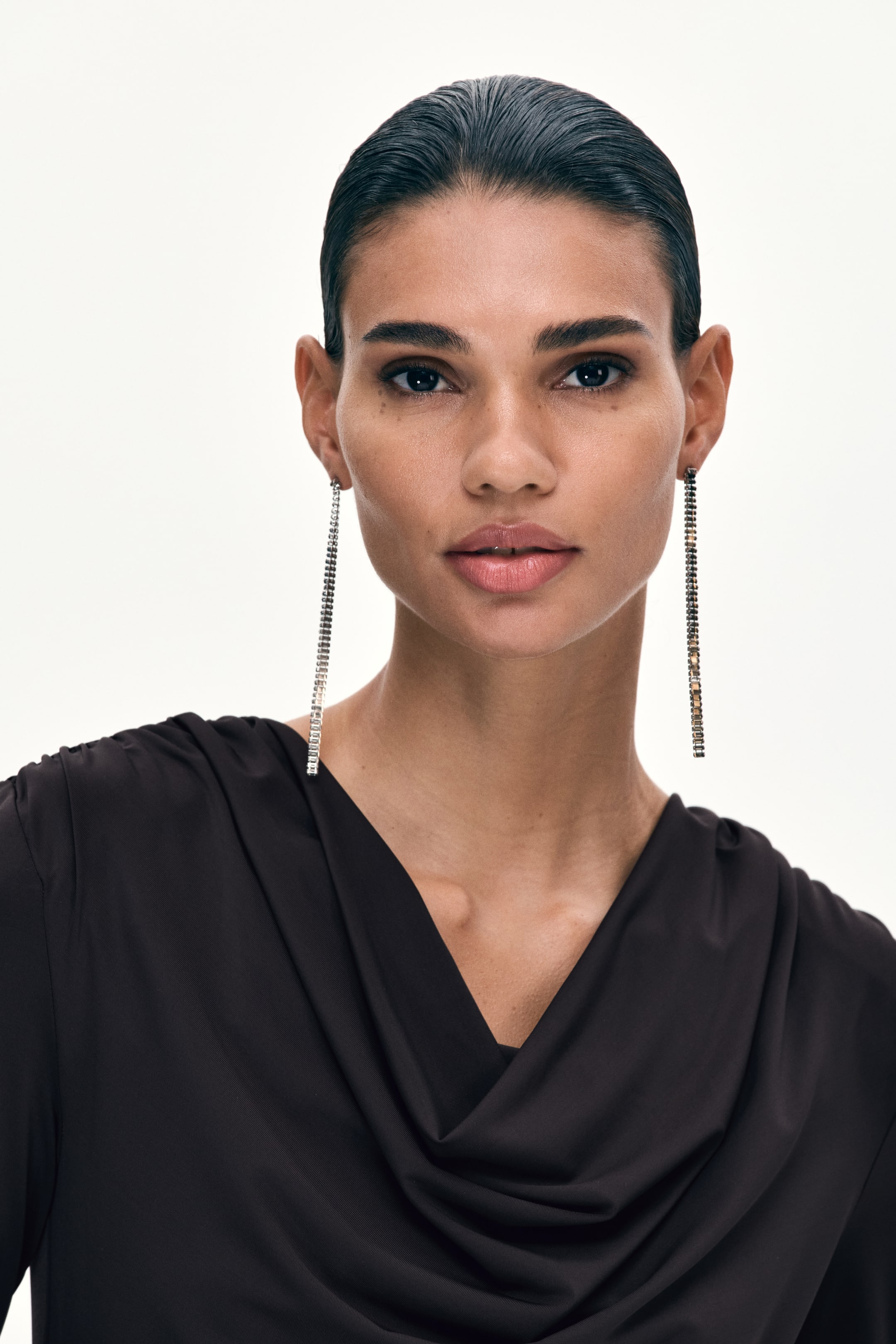 View larger image: A dark brown top featuring a draped cowl neck. Long, silver-toned dangle earrings sparkling with multiple rows of crystals.
