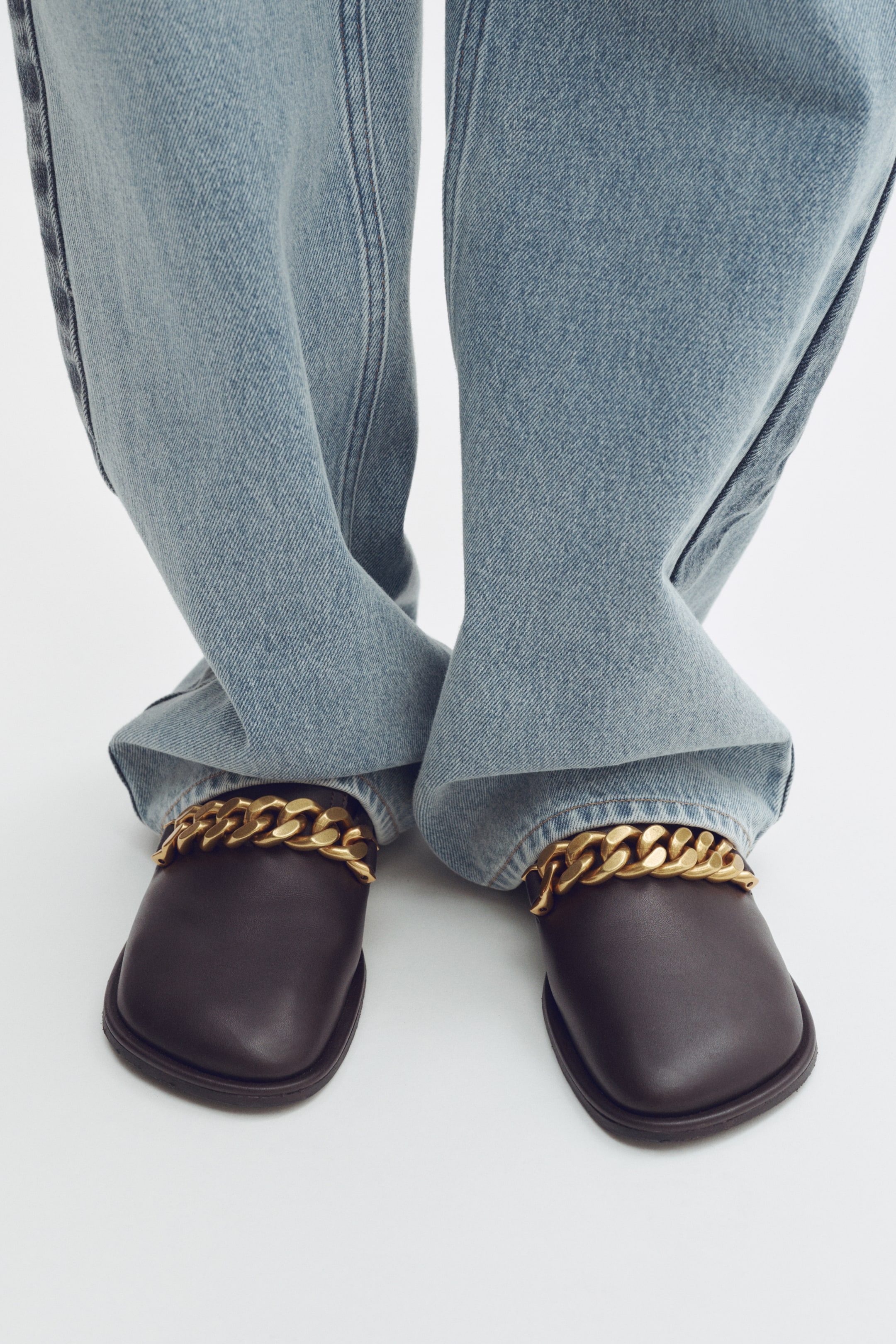 View larger image: Dark brown square-toe flats are showcased from a front-facing angle, each adorned with a prominent gold-toned chain detail across the vamp. The shoes are partially covered by light blue, wide-leg jeans, accentuating their design.
