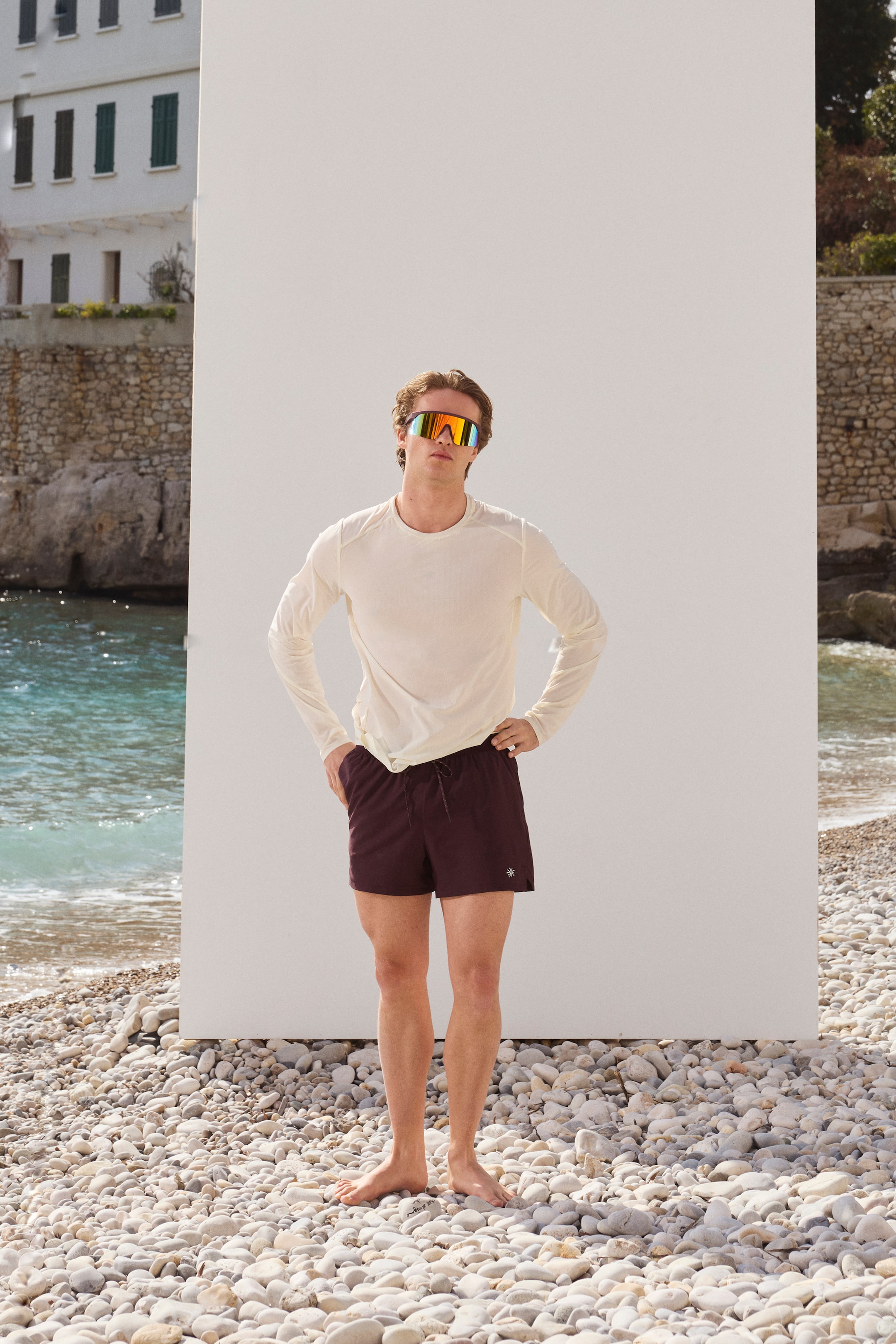 View larger image: Dark maroon swim shorts feature an elastic waistband with a visible drawstring and a small white emblem on the left leg. They are paired with a light cream crew neck long-sleeve shirt with raglan sleeves and multi-colored mirrored wrap-around sunglasses.