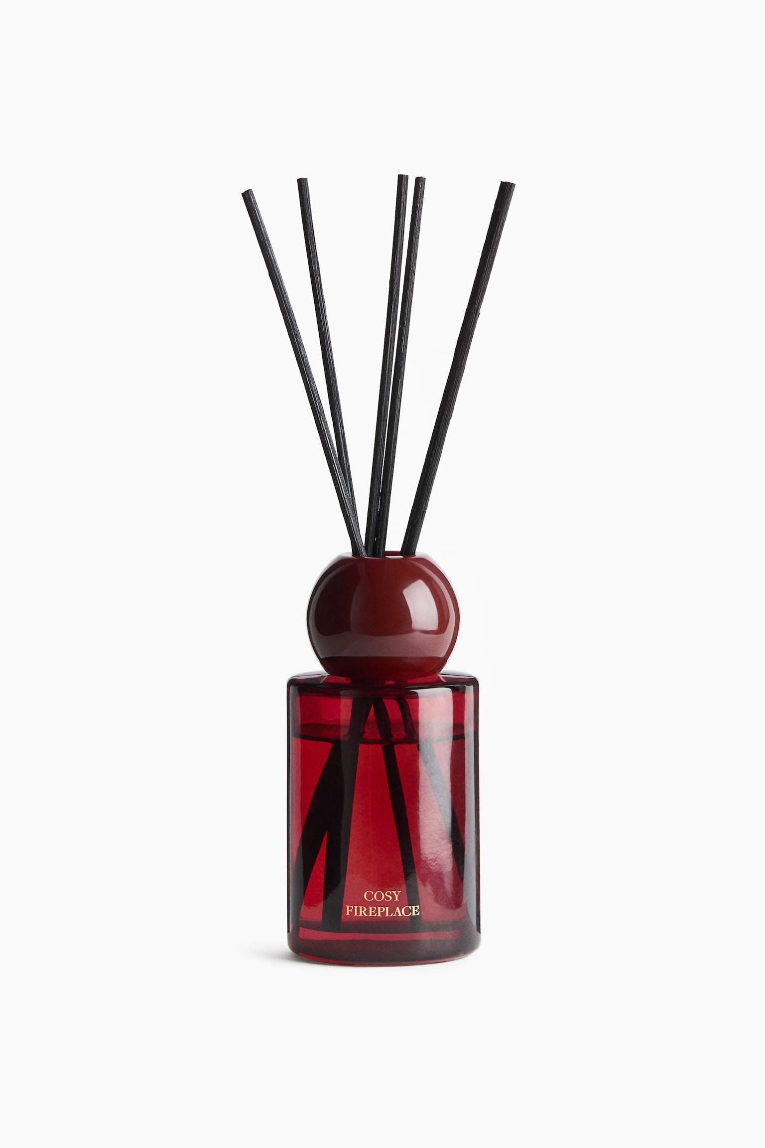 Una vista frontal muestra un difusor de varillas con una botella cilÃ­ndrica de vidrio rojo translÃºcido llena de aceite aromÃ¡tico. Cinco varillas de madera negras se abren en abanico desde un collar esfÃ©rico de vidrio rojo a juego en la parte superior, con las palabras "COSY FIREPLACE" impresas en dorado en la parte frontal de la botella.