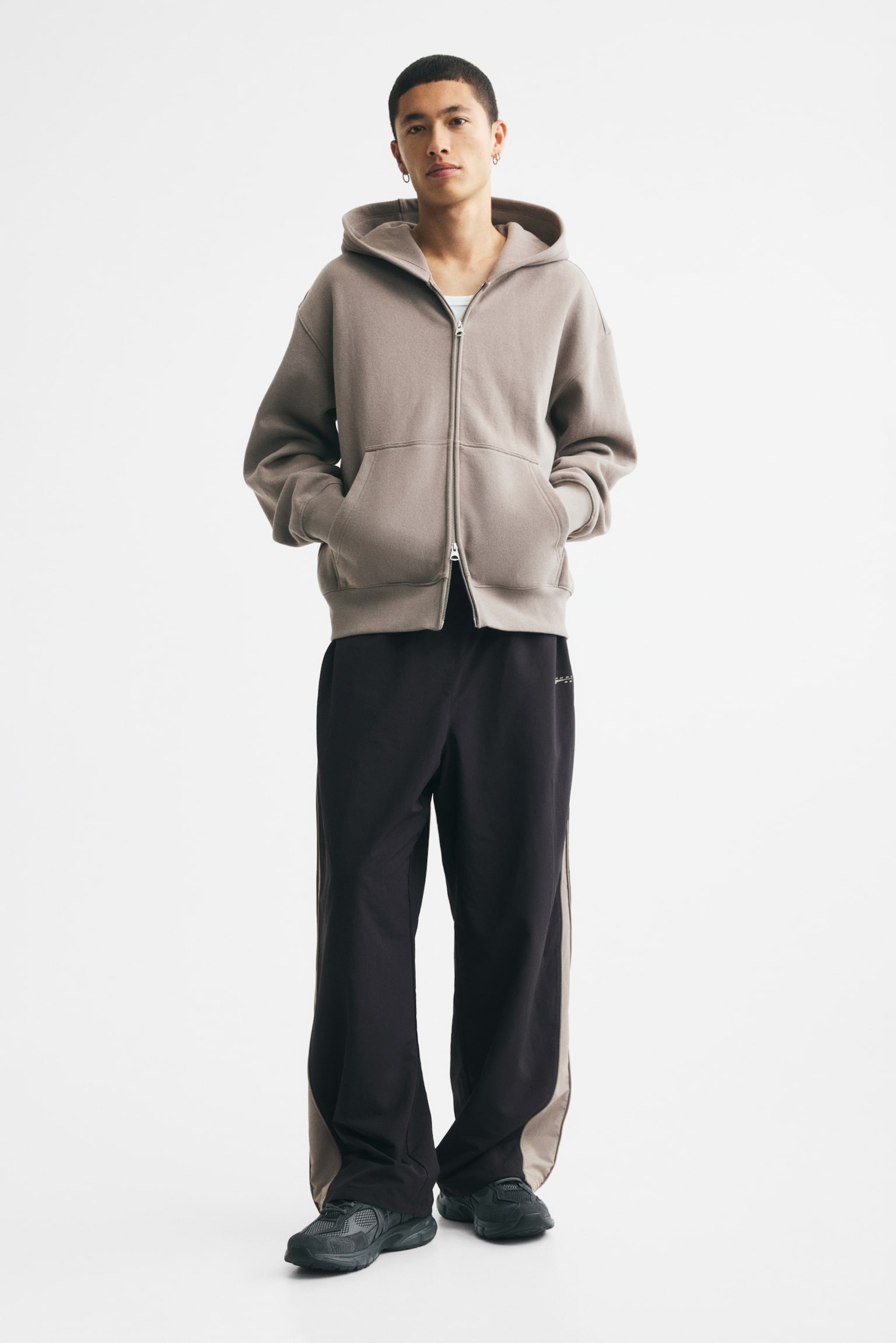 Black trousers are shown from the front, boasting an elasticated waistband and grey side panels extending down the outer seams of both legs. A small, light-coloured logo is visible on the upper left leg, and the elasticated leg hems feature adjustable drawstrings with toggles. - 1