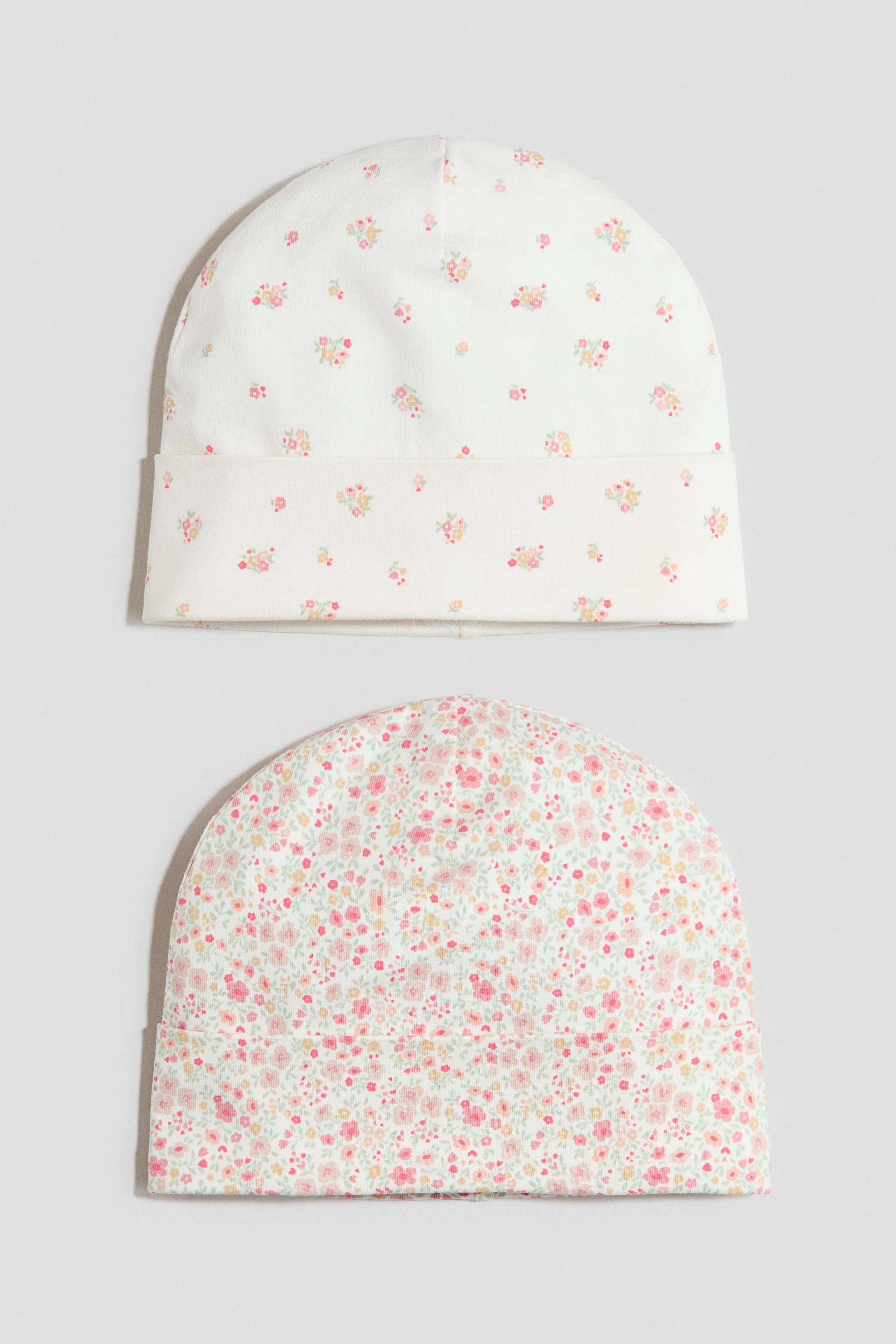 View larger image: Two soft cotton-jersey beanies are shown from the front. The top beanie has a cream base with a scattered pattern of small pink and yellow flowers and features a turn-up at the hem. The bottom beanie has a cream base with a dense pattern of small pink, yellow and green flowers and also features a turn-up at the hem.