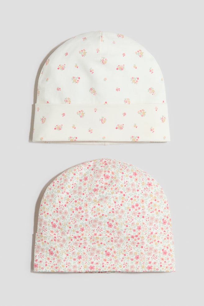 Two soft cotton-jersey beanies are shown from the front. The top beanie features a cream base with a scattered pattern of small pink and yellow flowers and has a turn-up at the hem. The bottom beanie features a cream base with a dense pattern of small pink, yellow and green flowers and also has a turn-up at the hem. - 1