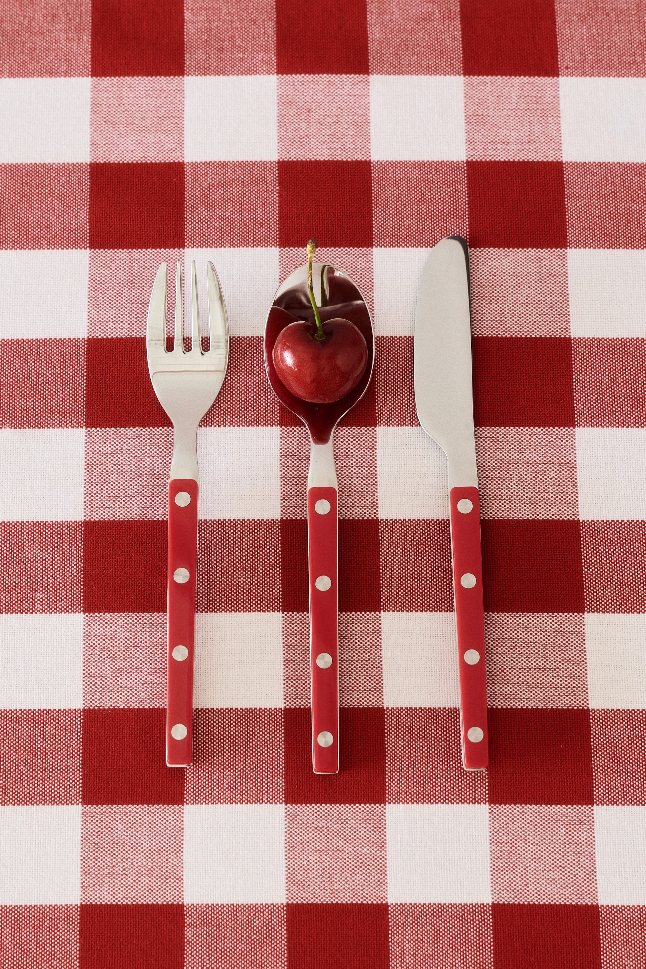 View larger image: A 3-piece children's cutlery set is laid flat, comprising a fork, a spoon, and a knife. Each piece is crafted from silver-coloured metal and features a red handle, adorned with three visible silver rivets.