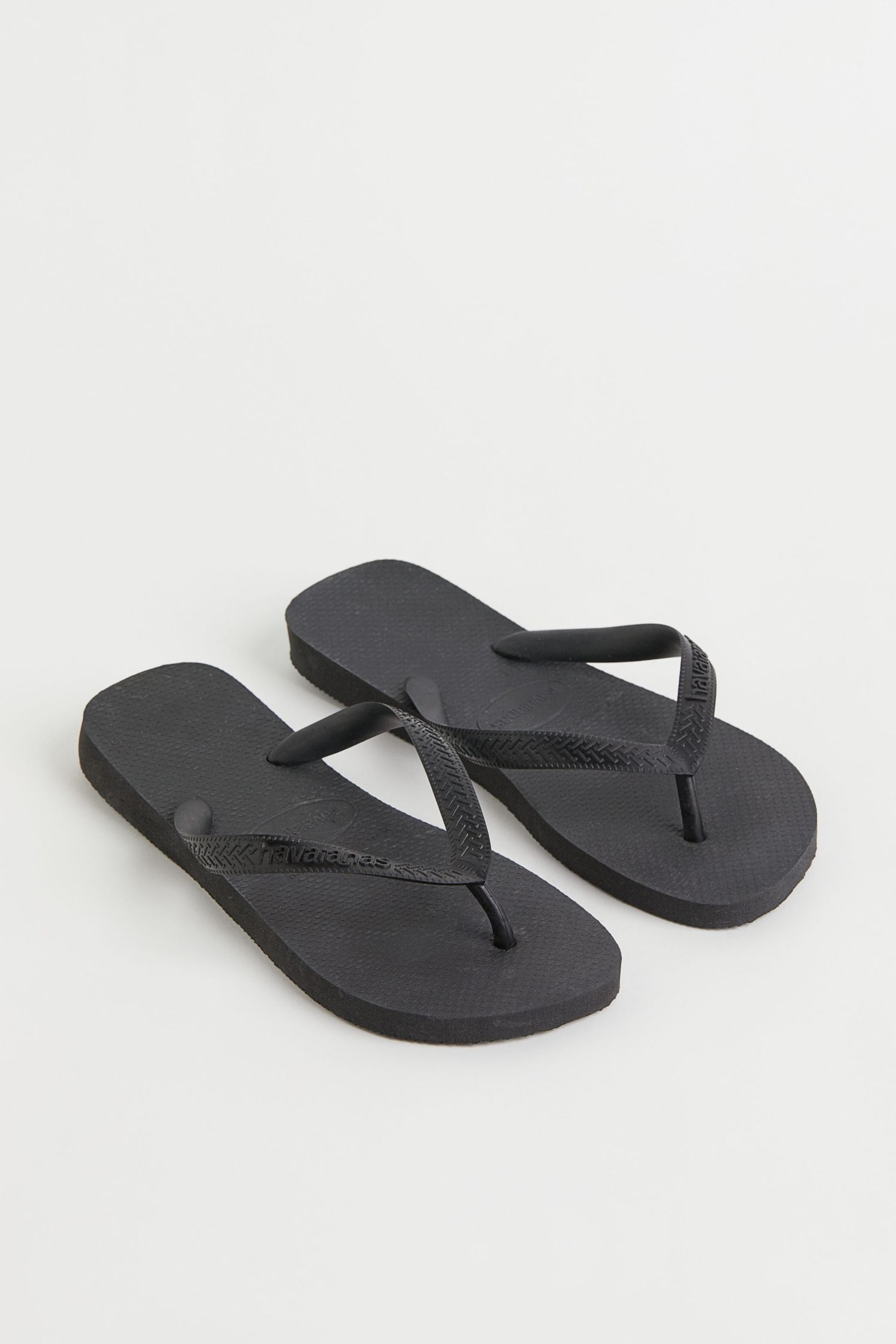 Visa större bild: Ett par svarta flip-flops presenteras i en vinkel, med tårna mot övre vänstra hörnet. Varje flip-flop har en strukturerad sula och V-formade remmar, som visar ett chevronmönster och varumärket "havaianas" på en av remmarna.