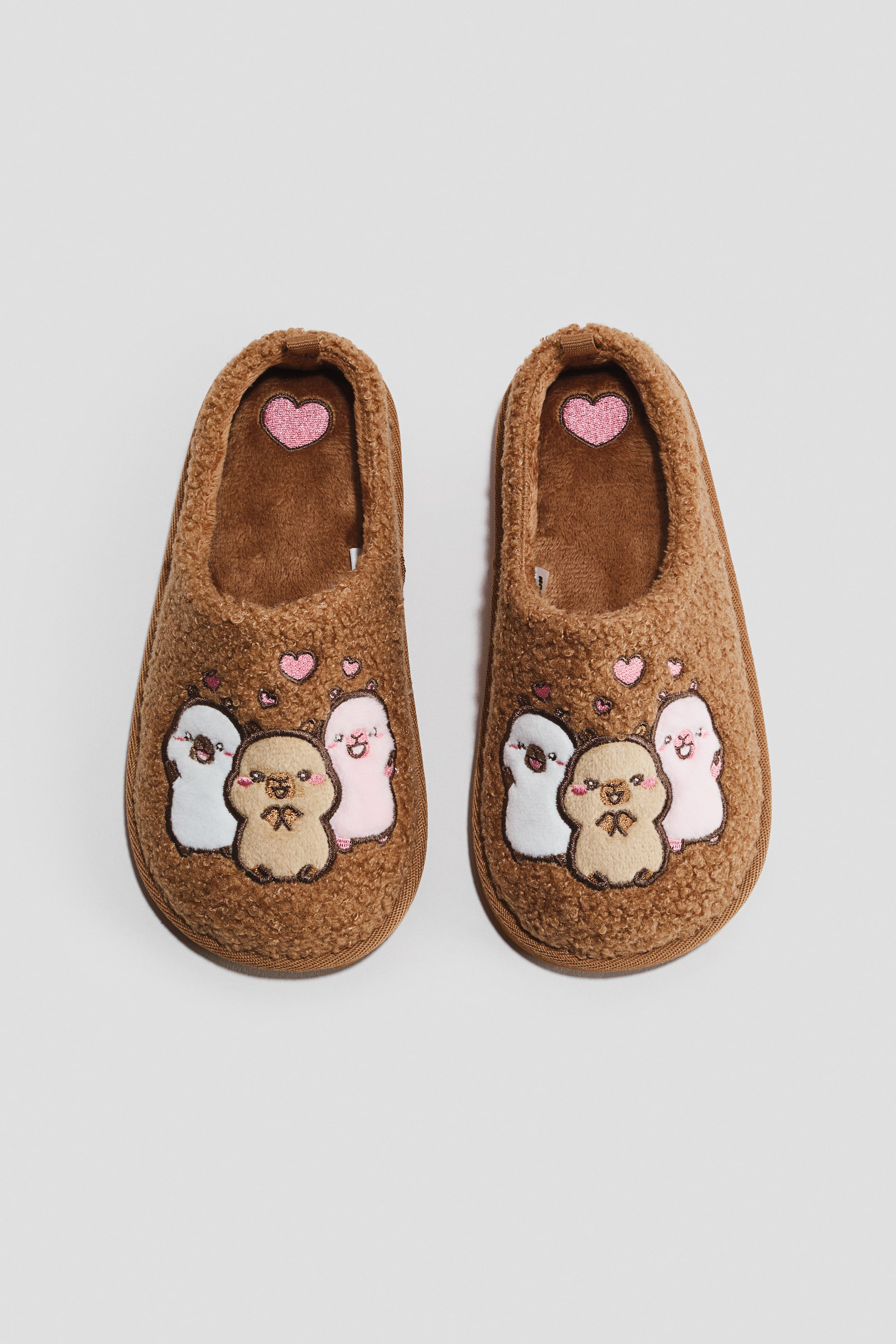 View larger image: Embroidered-motif teddy slippers feature a brown, fuzzy texture and an embroidered design across the top, viewed from a top-down angle. The design showcases three cartoon animal characters: a centrally positioned brown one, a white one on the left, and a pink one on the right, with small pink hearts floating above them. Additionally, a pink glitter heart details the heel of each plush inner sole.