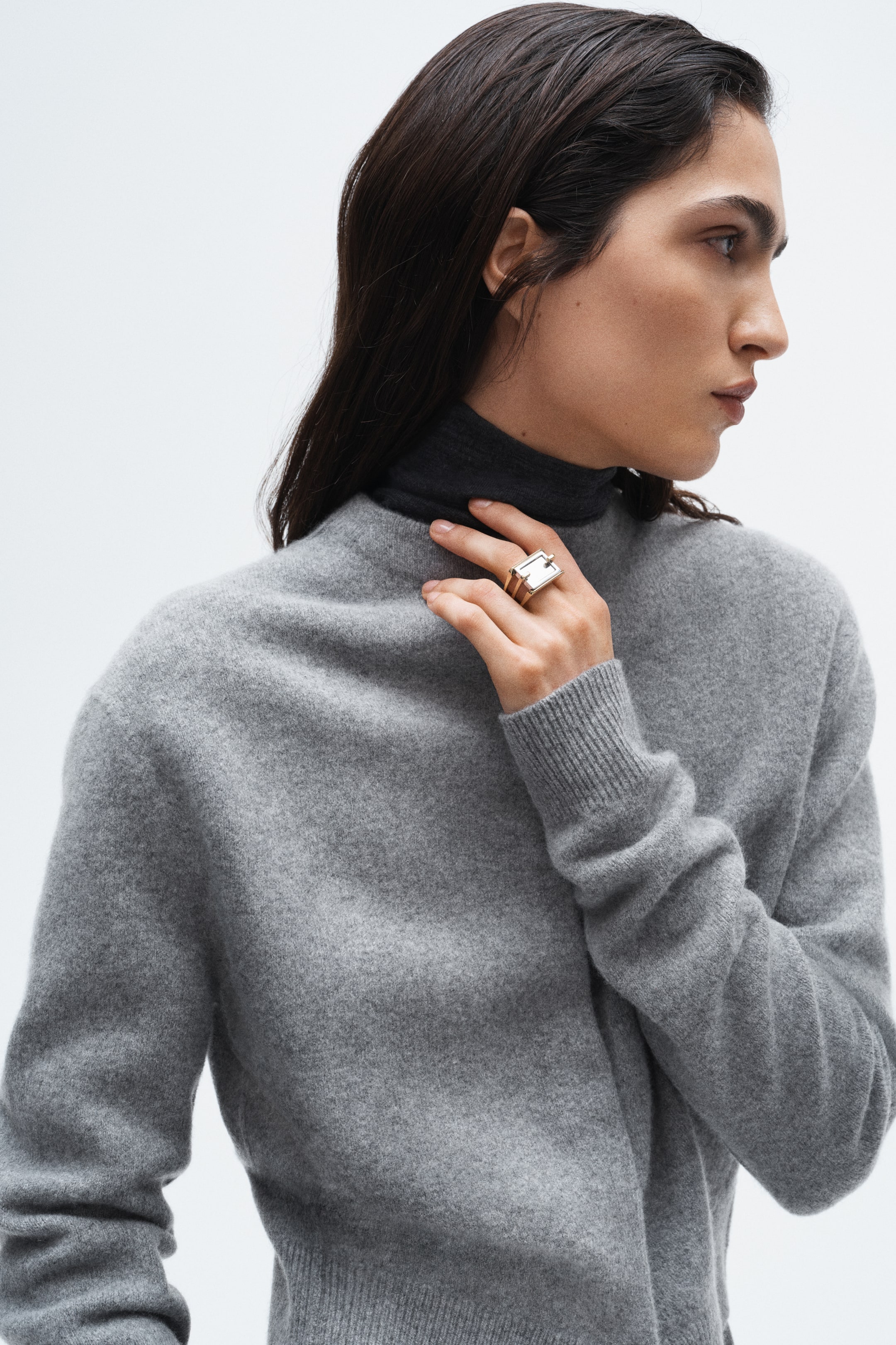 View larger image: A gold-toned ring featuring a rectangular, translucent stone is being worn on the model’s left ring finger, while the model’s left hand is touching a dark grey turtleneck at their neck.