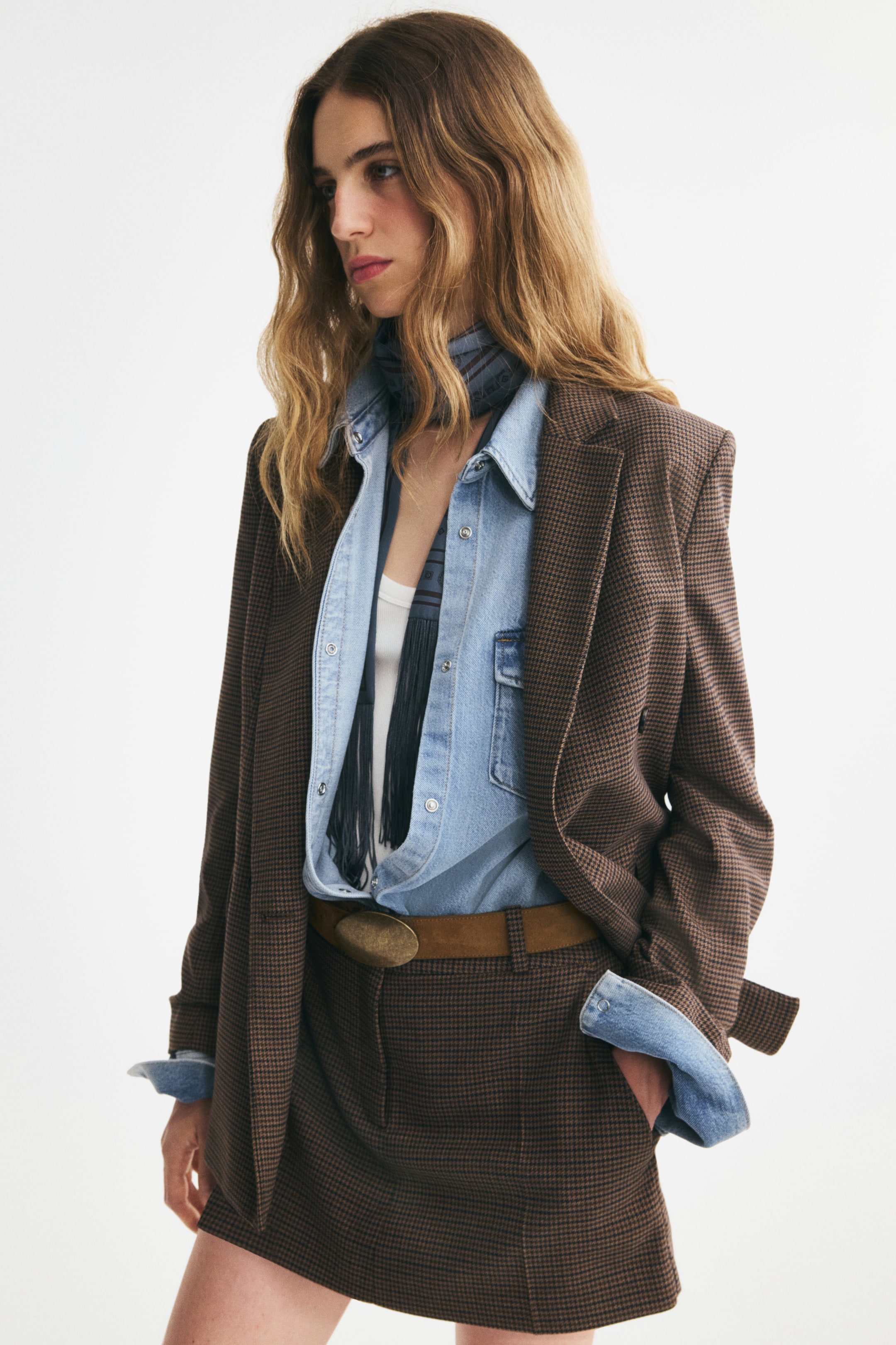 View larger image: A brown houndstooth, double-breasted blazer in a slightly longer style is worn open from a three-quarter front angle, highlighting its structured shoulders and notch lapels. The blazer is layered over a light-blue denim shirt with folded-back cuffs and paired with a matching houndstooth skirt.