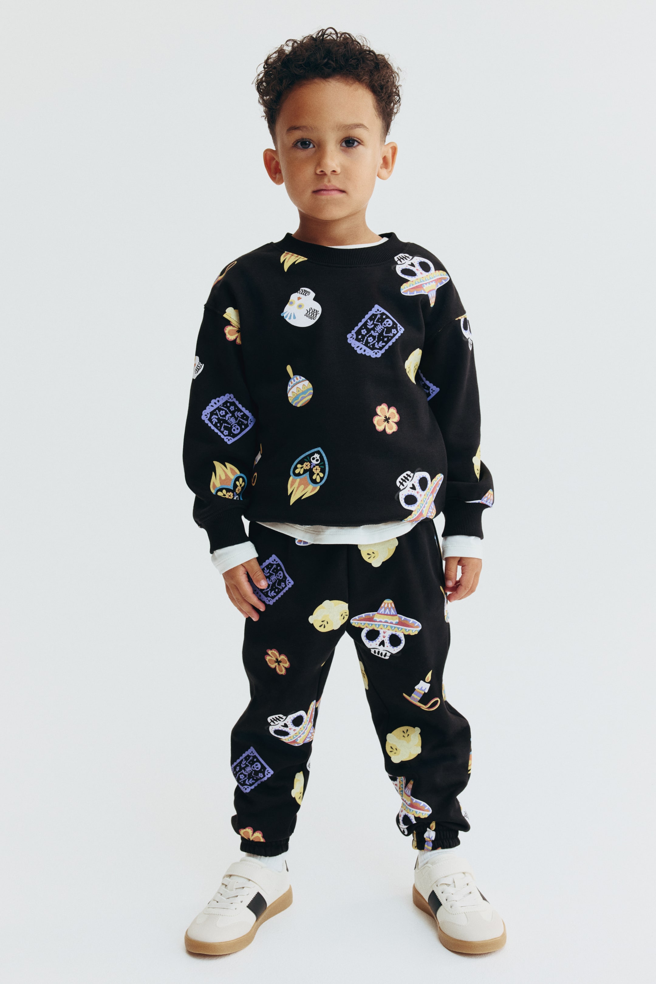 View larger image: A black matching sweater and track pants set is shown from the front, featuring an all-over colorful print of cartoon skulls, flowers, and festive icons. The long-sleeved sweater is designed with a round ribbed neckline, dropped shoulders, and ribbed cuffs and hem, while the matching track pants feature an elasticated waistband and elasticated hems.