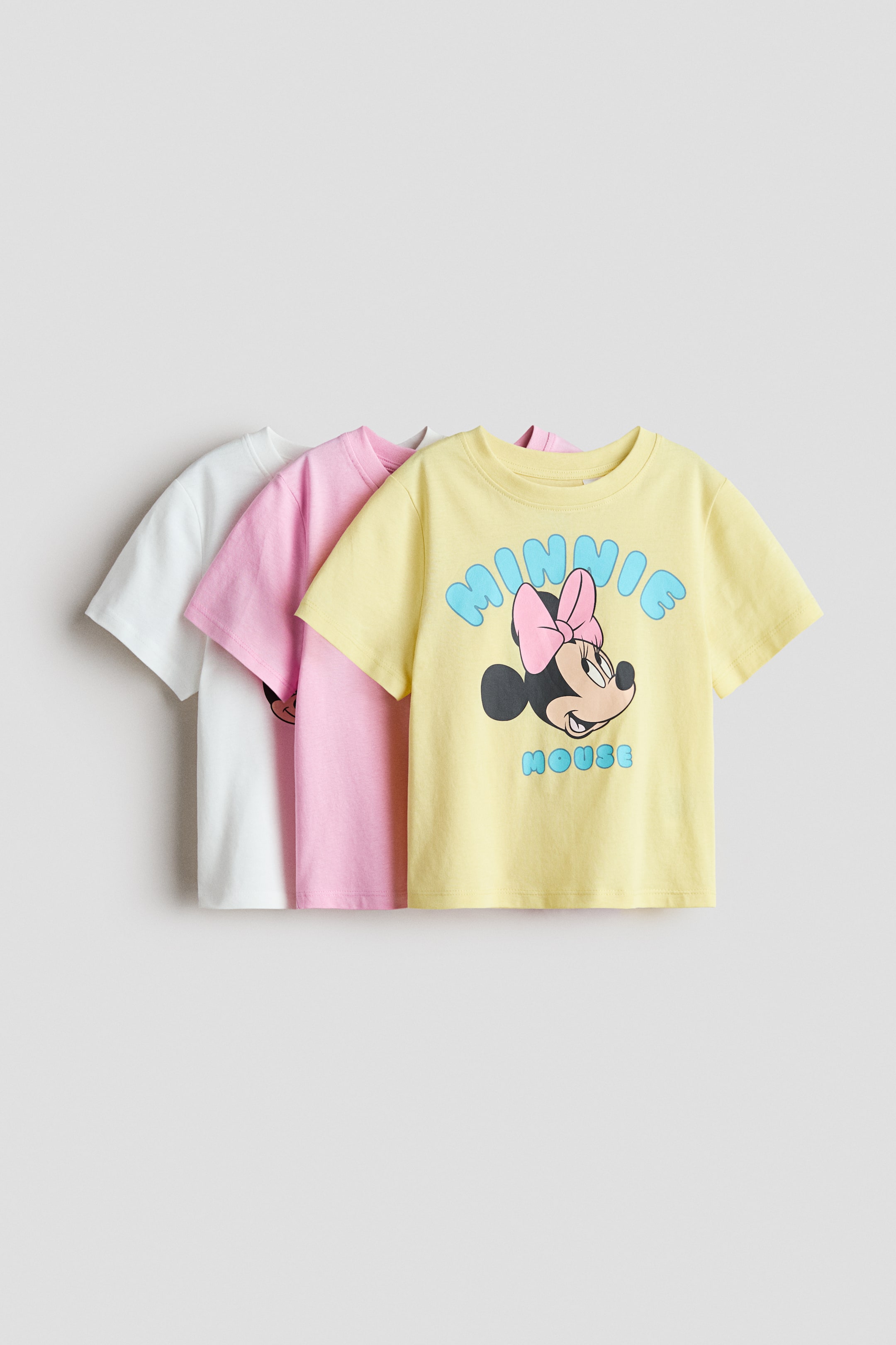 View larger image: Tatlong short-sleeved na tops na gawa sa malambot na cotton jersey ay nakaayos, bahagyang magkapatong, nakaharap sa harap. Ipinapakita ang mga ito sa puti, pink, at dilaw, kung saan ang dilaw na top ay mayroong Minnie Mouse print motif.