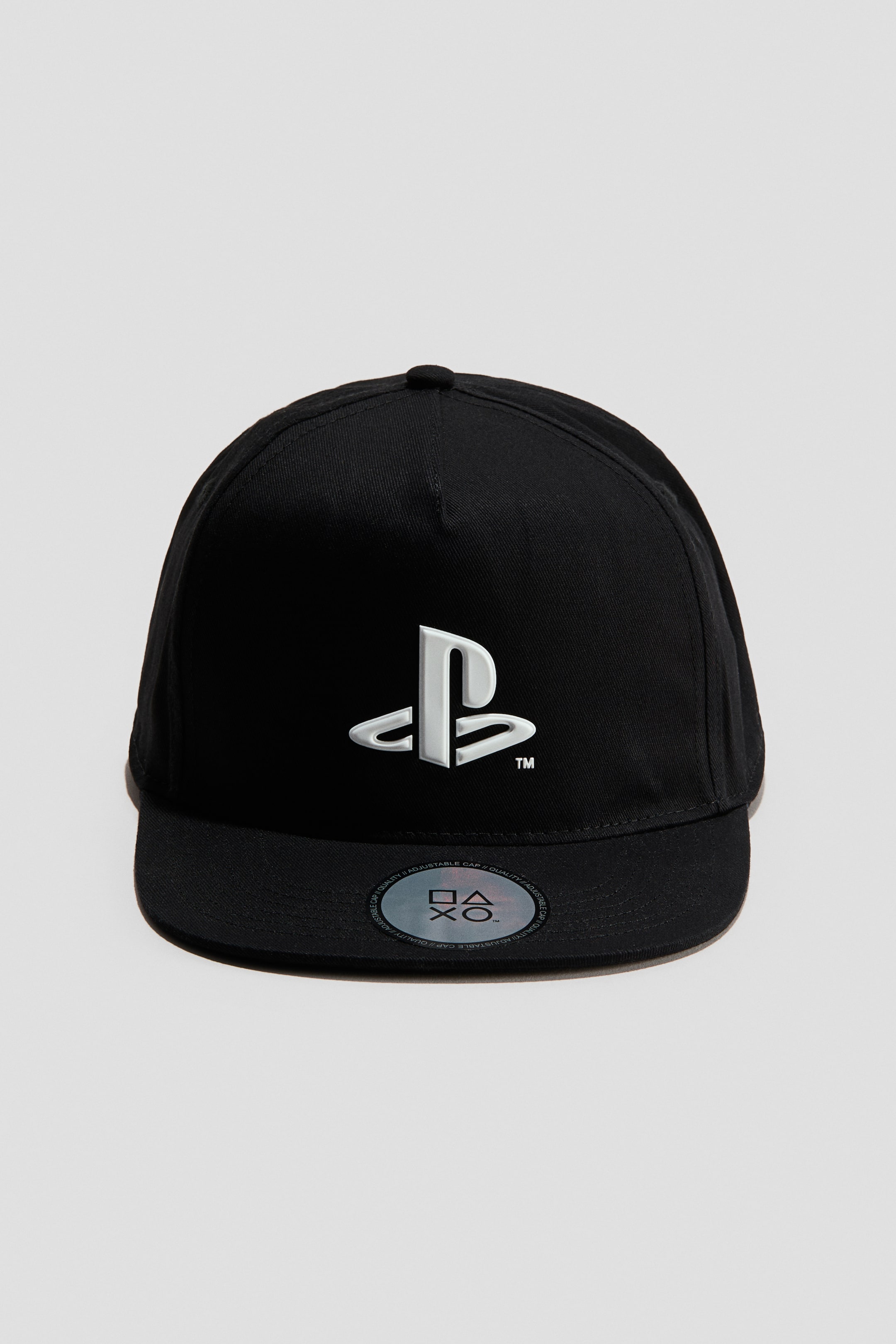 View larger image: A black cotton twill cap is facing forwards, displaying a white PlayStation logo printed on the front panel. The cap features a flat peak with a circular holographic sticker attached to the top.