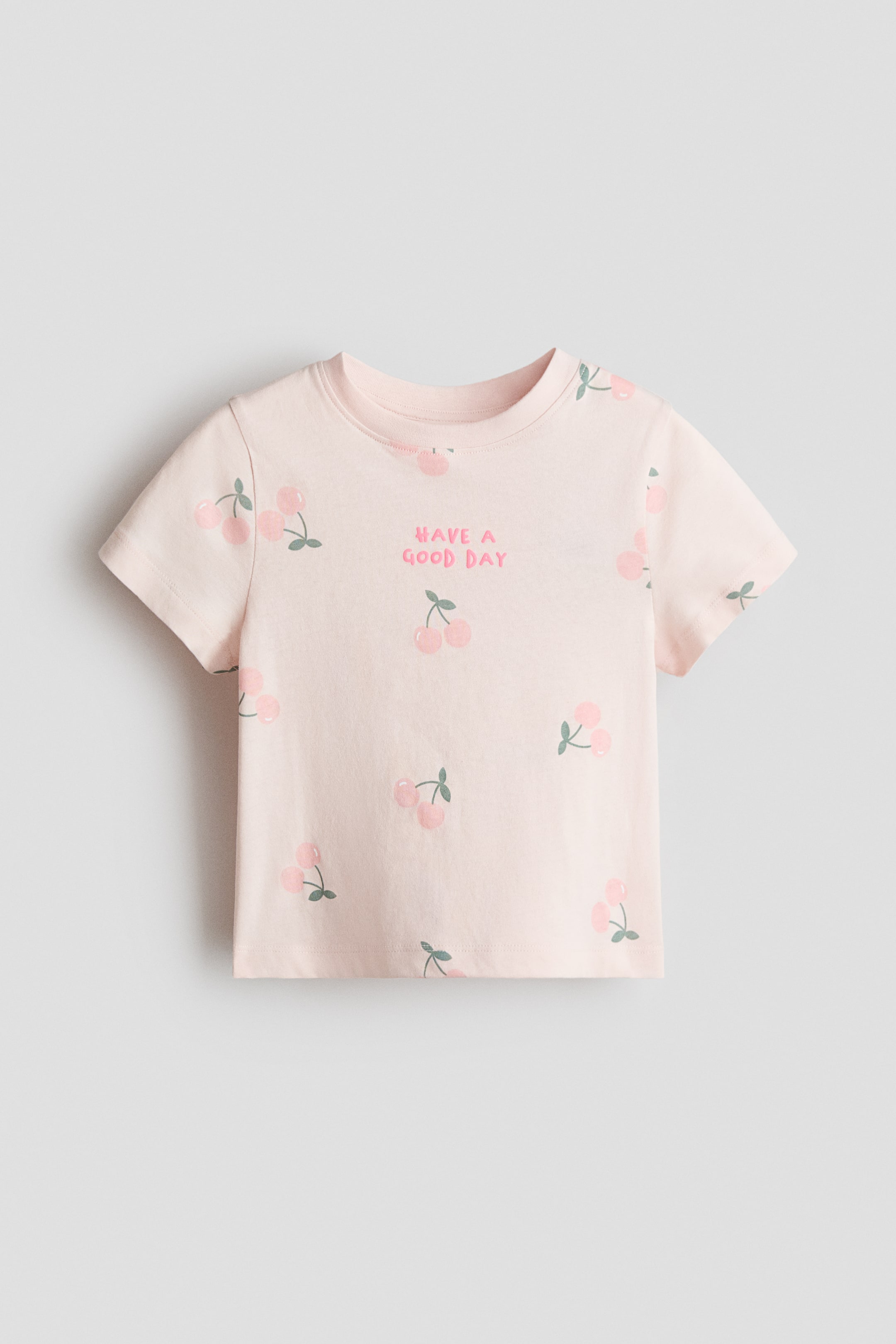 View larger image: A light pink T-shirt is presented FRONT, featuring a round neckline and short sleeves. It is adorned with a scattered cherry pattern and the phrase 'HAVE A GOOD DAY' in pink lettering.