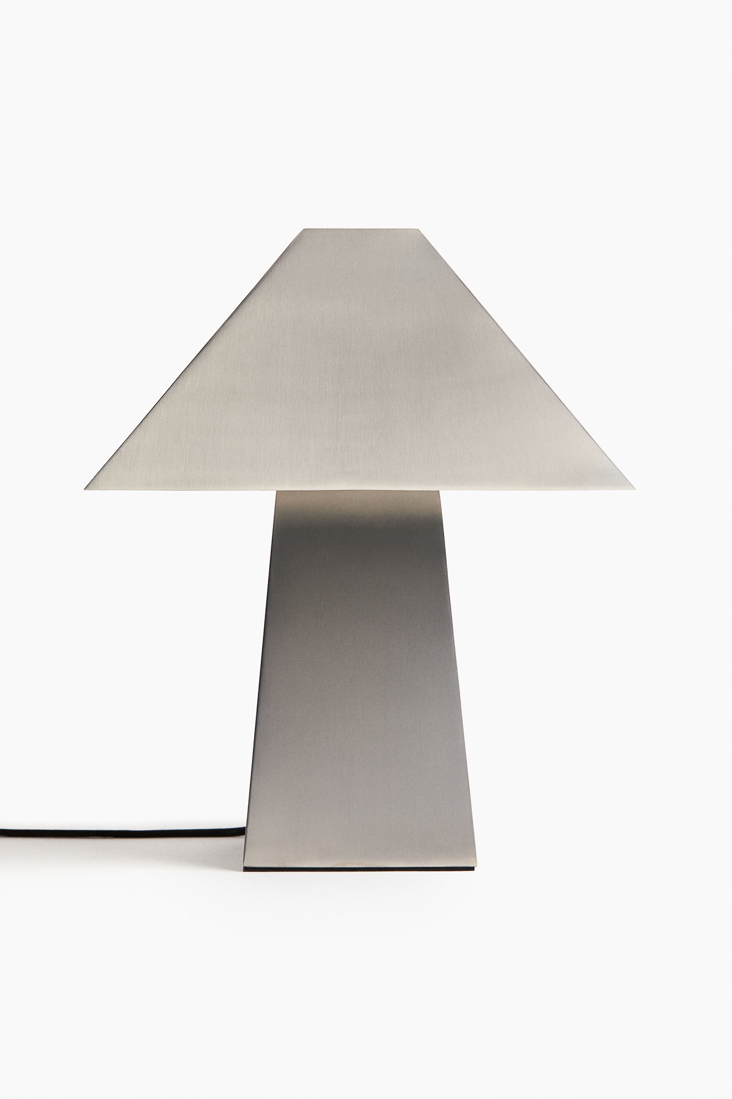 A metal table lamp with a geometric design is shown facing forwards, featuring a trapezoidal shade resting on a tapering rectangular base with a brushed finish. The lamp emits a warm downward glow from beneath the shade, illuminating the upper portion of the base.