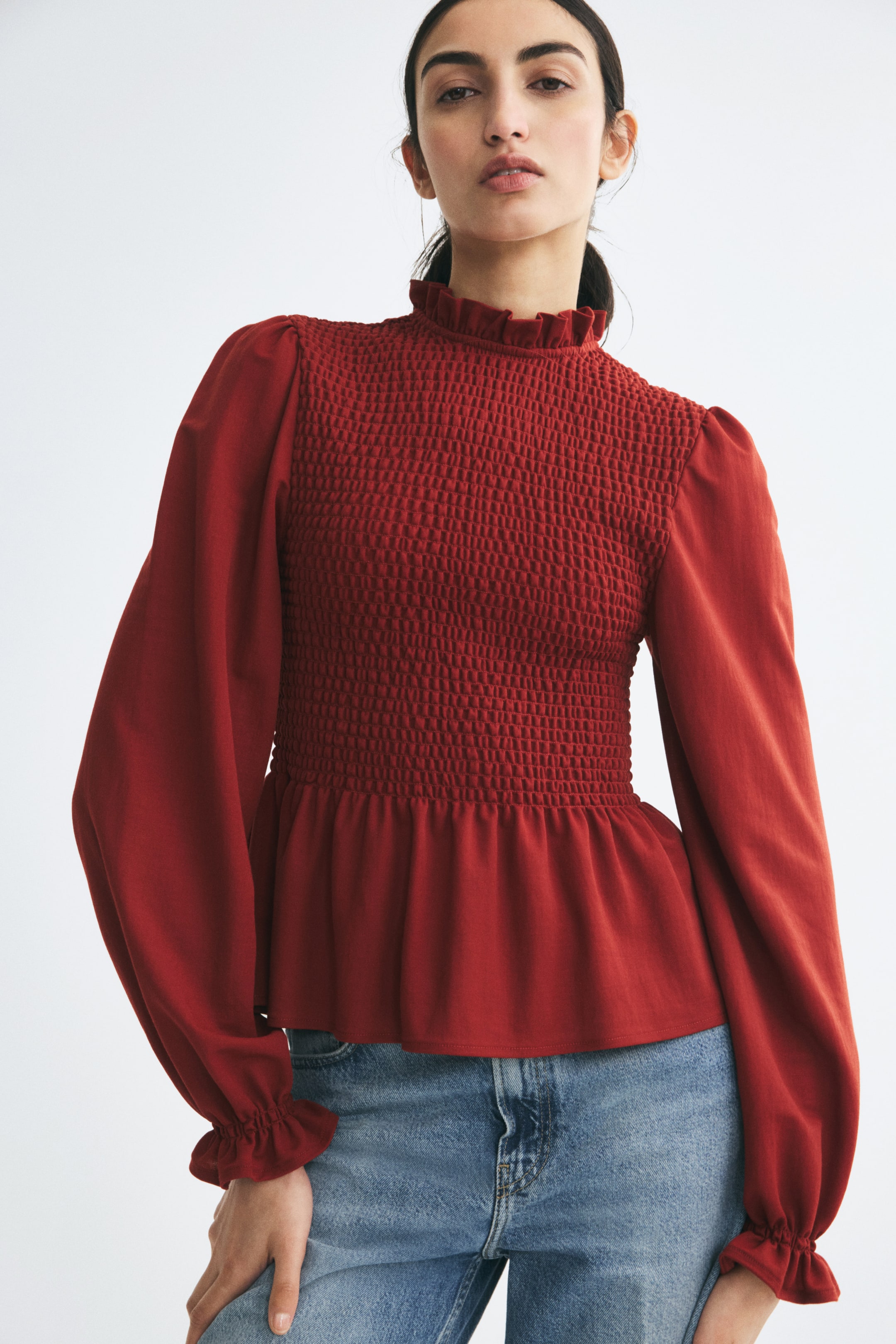 View larger image: A rust-red fitted top featuring a smocked bodice, a high frill collar, and a flared peplum hem is being worn by the model. The top is styled with light-blue jeans and features long puff sleeves with elasticated, ruffled cuffs.