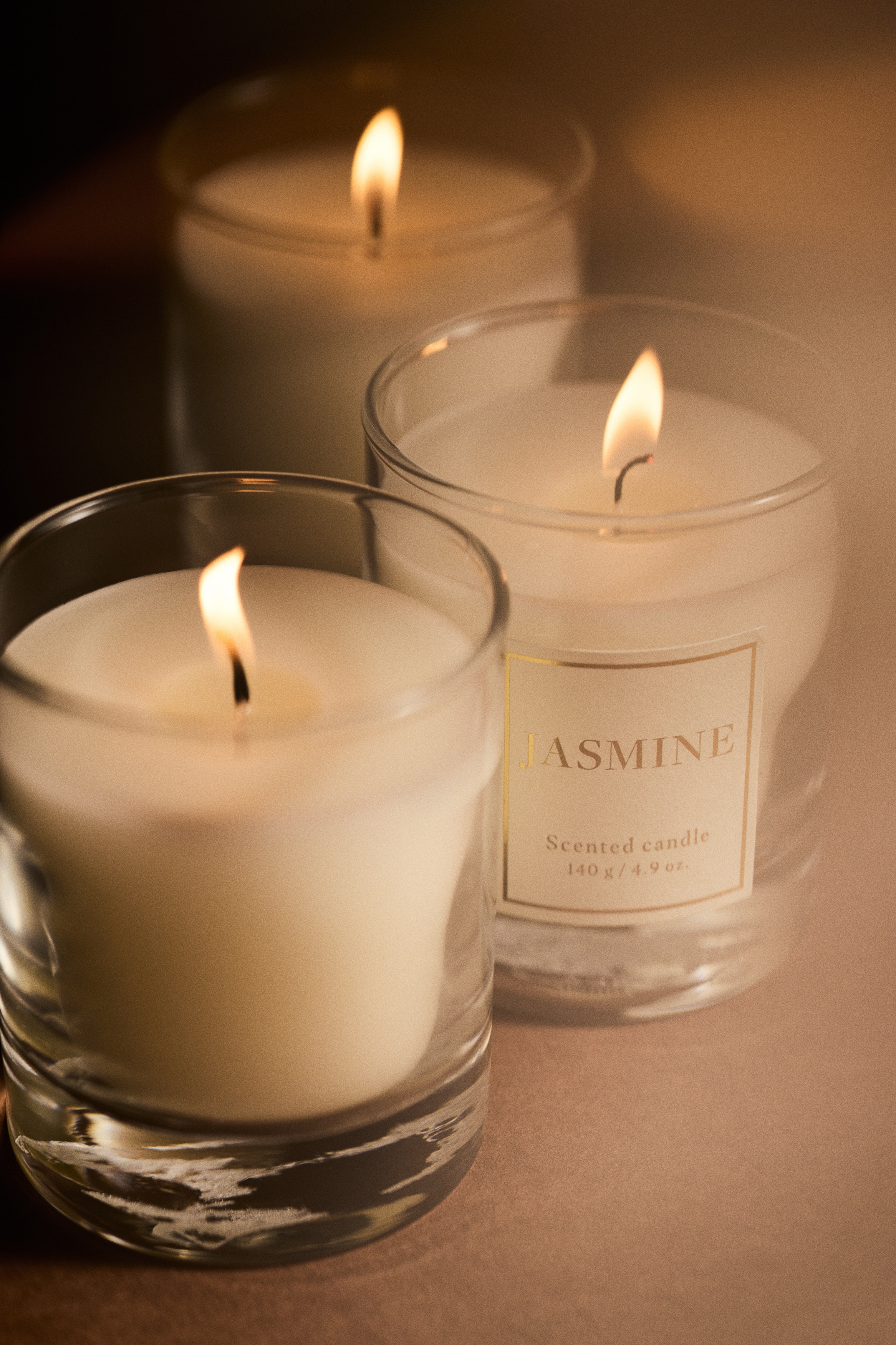 A white scented candle in a clear glass holder with a thick base is standing next to its matching square gift box, both facing front. The candle and the white box, which is decorated with a red dotted floral pattern, both feature a central white label with gold borders and lettering that reads "JASMINE".