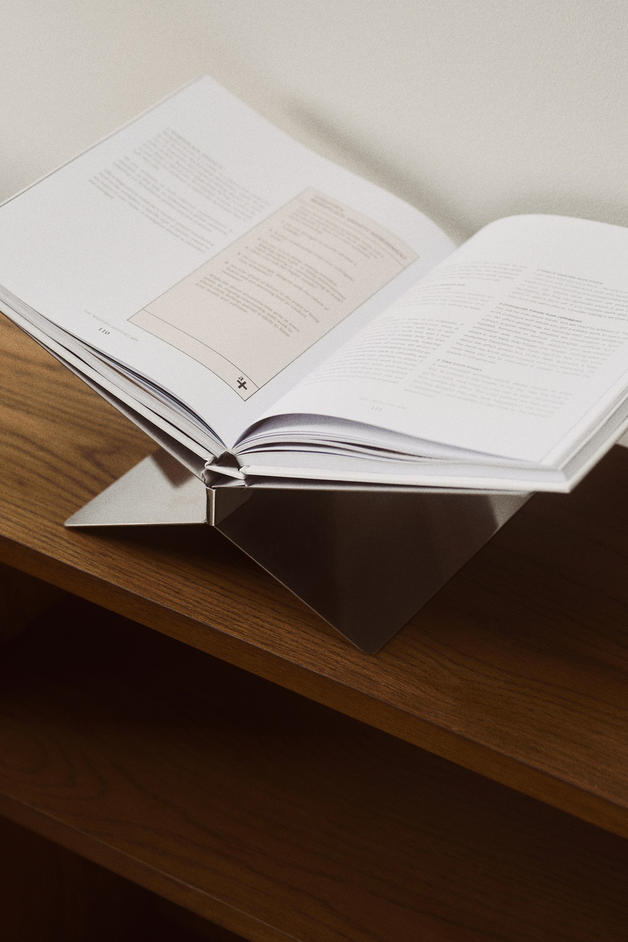 Voir une image plus grande: Un support de livre en métal maintient un livre ouvert, arborant une finition polie de ton argenté et un design angulaire et plié qui soutient les pages du livre.