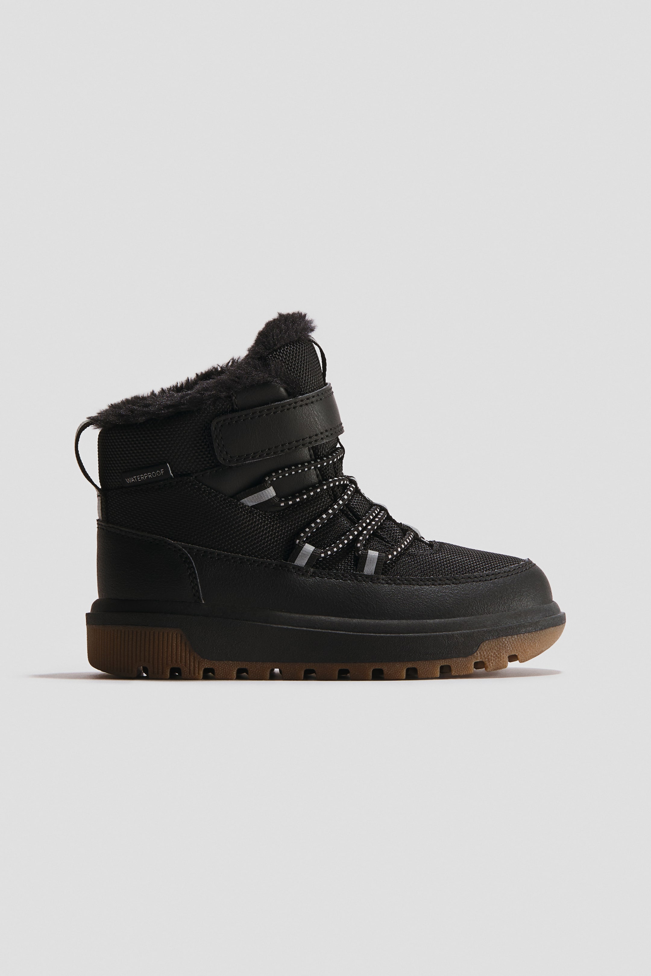 Se større billede: En sort vandtæt hi-top sneaker præsenterer sin side-front profil, med en sort overdel og imiteret pelsforing ved kraven. Den fremviser reflekterende snørebånd, en velcrorem over anklen, "WATERPROOF" tekst på siden og en kraftig, gummifarvet sål med et synligt slidmønster.