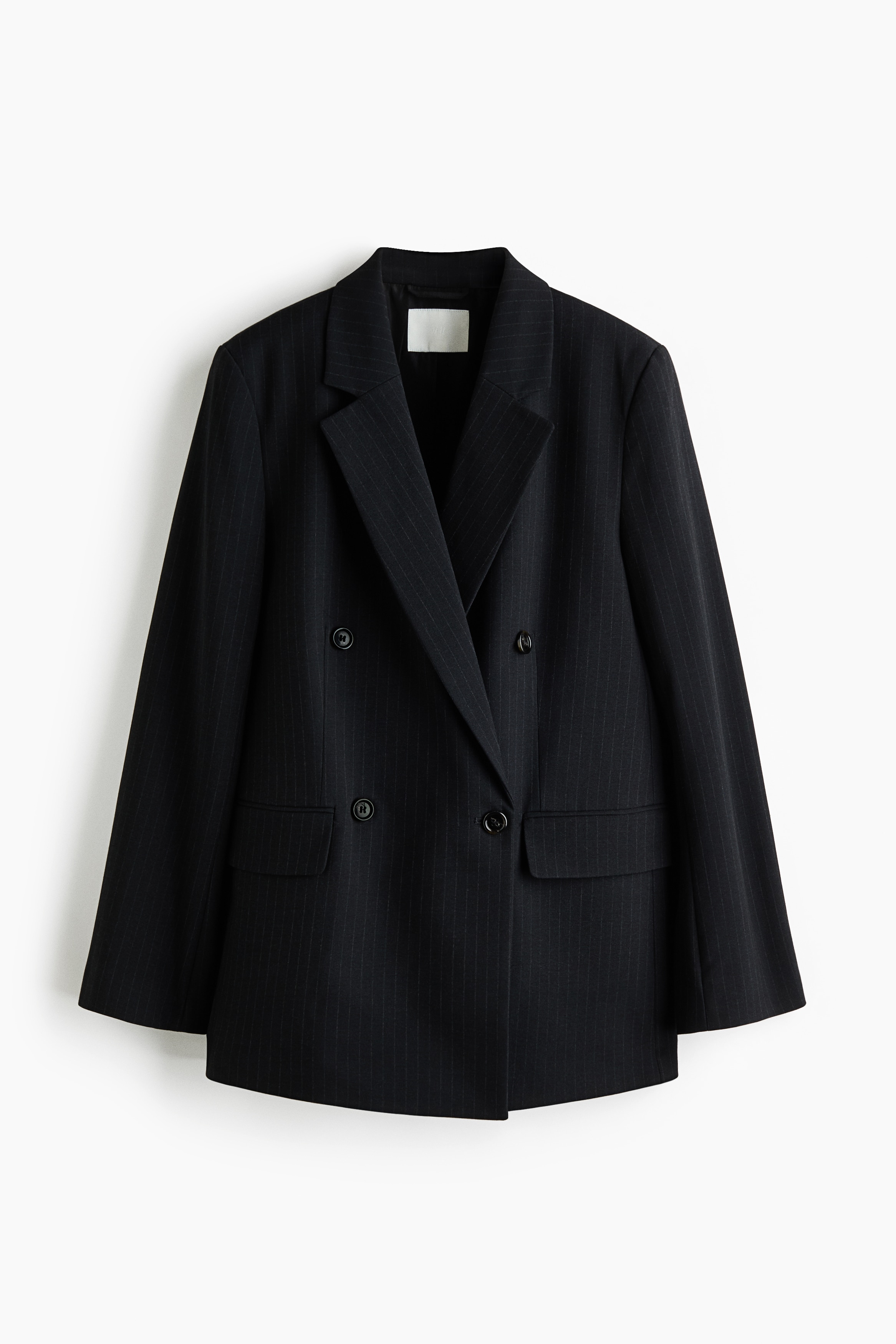 En sort dobbeltradet blazer med smalle striber bÃ¦res Ã¥bent af modellen, lagt over en hvid skjorte uden knapper og sorte bukser. Blazeren har markerede skulderpuder og revers med hak, og Ã¦rmerne er skubbet op for at vise de opsmÃ¸gede hvide skjortemanchetter.