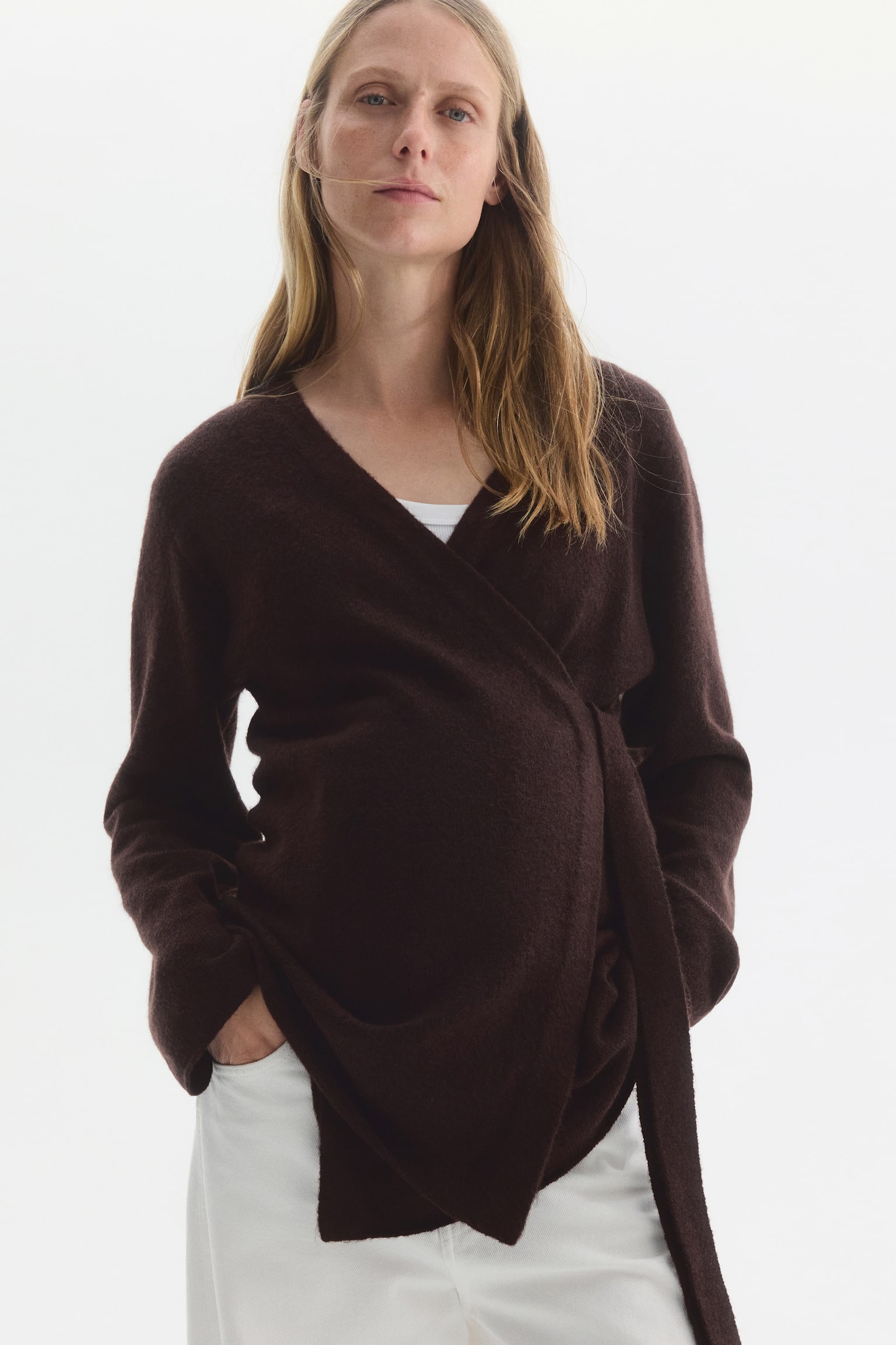 The model is wearing a dark brown fine-knit wrapover cardigan over a white top and white trousers. The cardigan features a wrap front with a V-neckline, dropped shoulders, and a long fabric tie hanging to one side. - 1