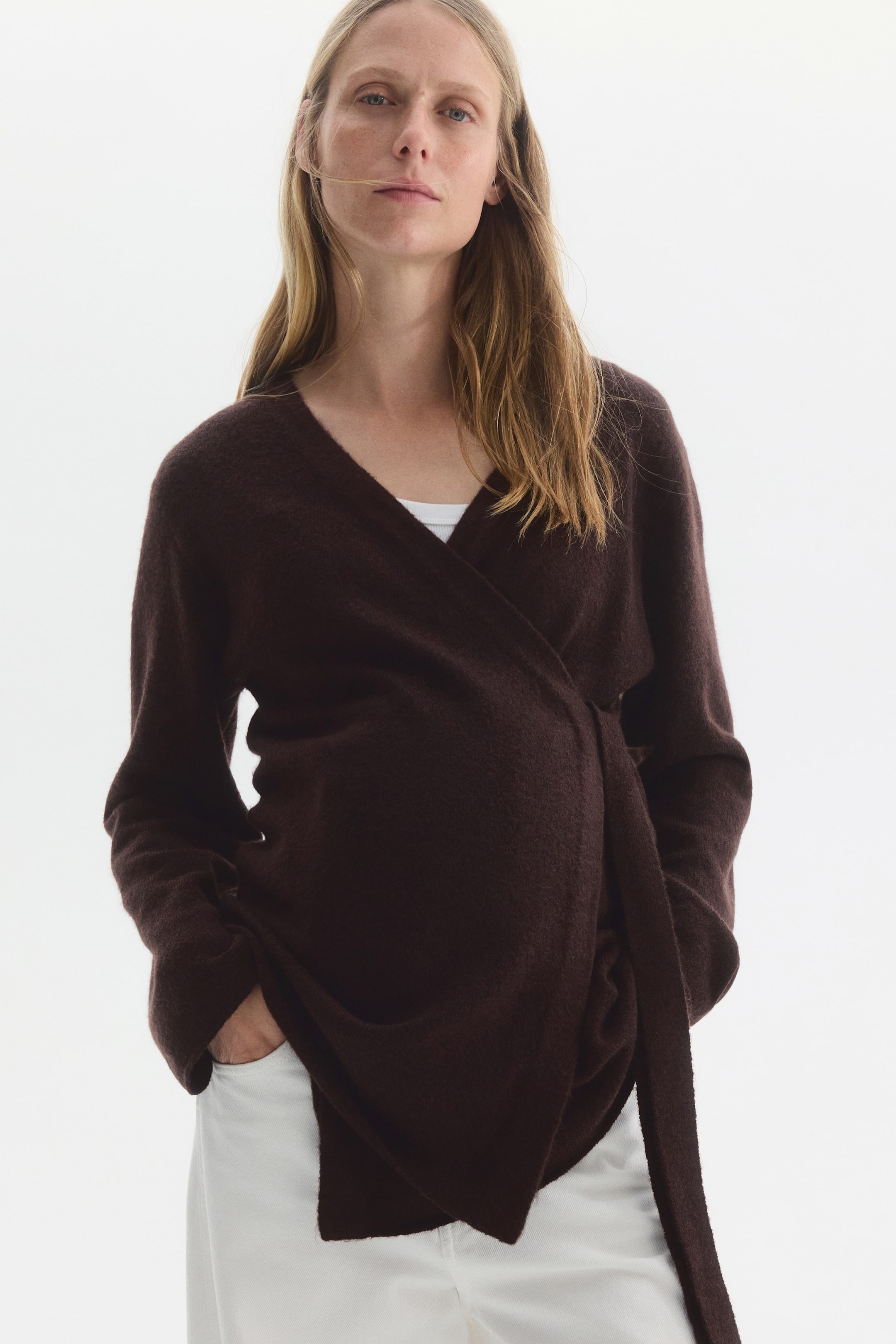 Visualizza l'immagine ingrandita: Un cardigan avvolgente in maglia fine di colore marrone scuro è indossato dalla modella sopra un top bianco e pantaloni bianchi. Il cardigan presenta una chiusura incrociata sul davanti con scollo a V, spalle scese e un lungo laccetto in tessuto che pende su un lato.