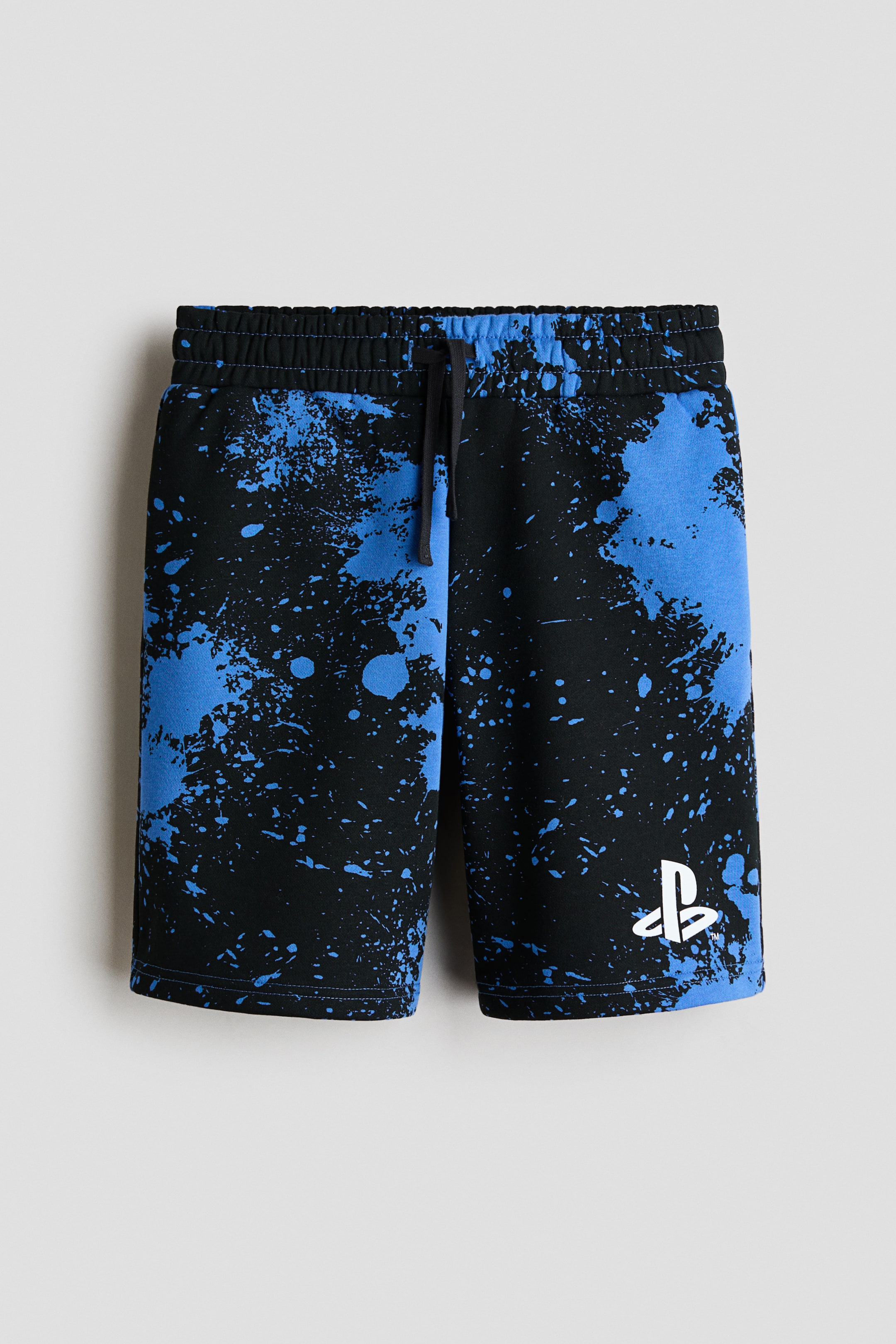 Ampliar la imagen: Se muestra un par de shorts de felpa hasta la rodilla desde el frente, con un estampado de salpicaduras de pintura en negro y azul brillante. Los shorts lucen una cintura elástica con cordón negro y un logotipo blanco de PlayStation impreso en la parte inferior de la pierna izquierda.