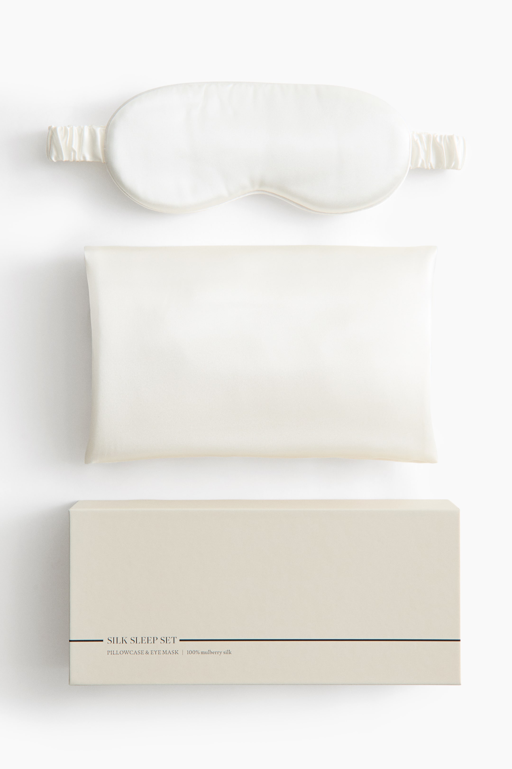 View larger image: A silk sleep set is displayed from the front in a flat-lay view, featuring an off-white padded sleep mask with a gathered elastic strap and a matching rectangular silk pillowcase. Below them, a beige rectangular gift box is shown with the printed text "SILK SLEEP SET" along the bottom edge.