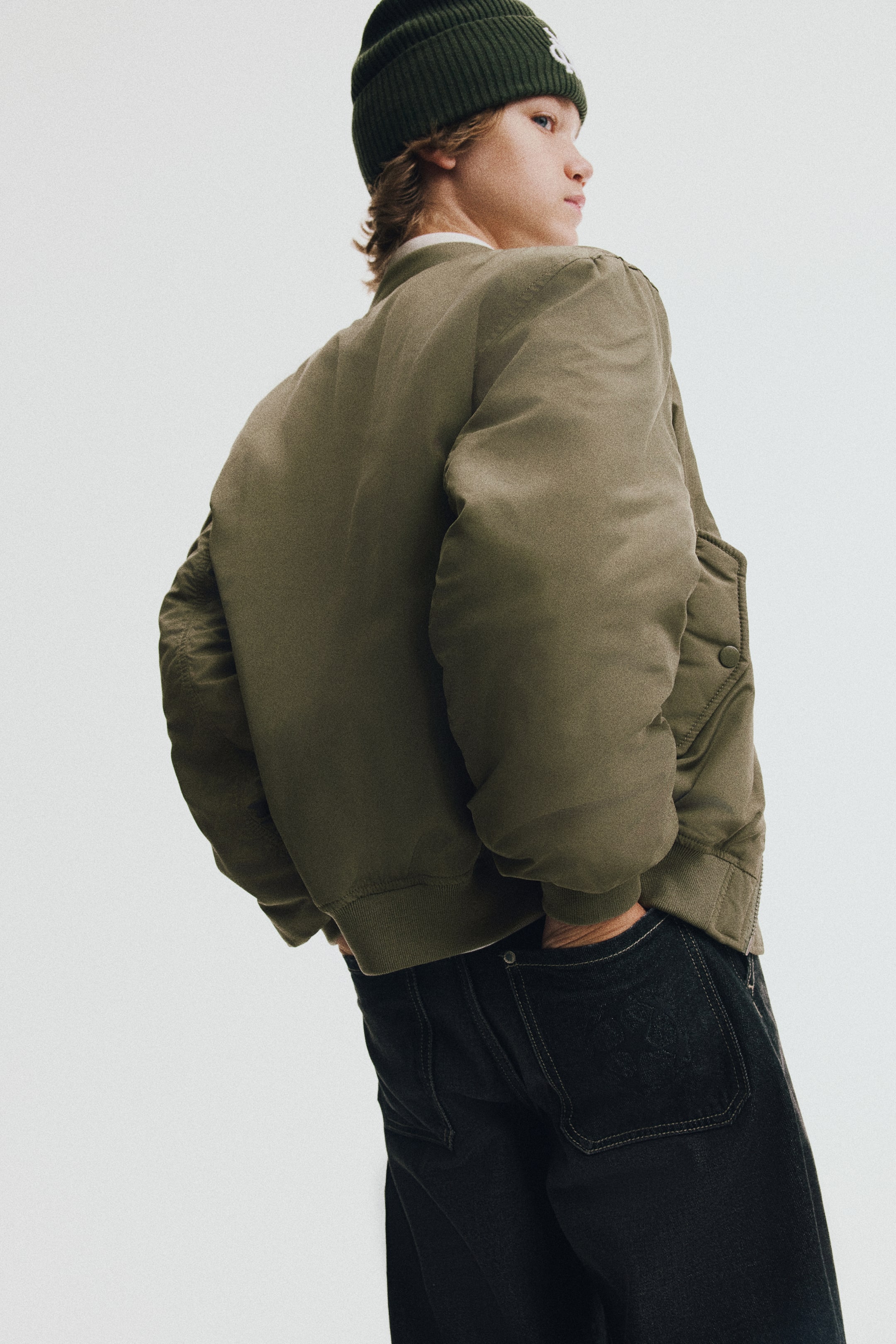 View larger image: A lightly padded olive green bomber jacket is shown from a back three-quarter angle, highlighting its relaxed fit, dropped shoulders, and ribbed hem. The model is wearing the jacket paired with dark jeans and a green beanie, standing with hands in the back pockets to showcase the garment’s drape.
