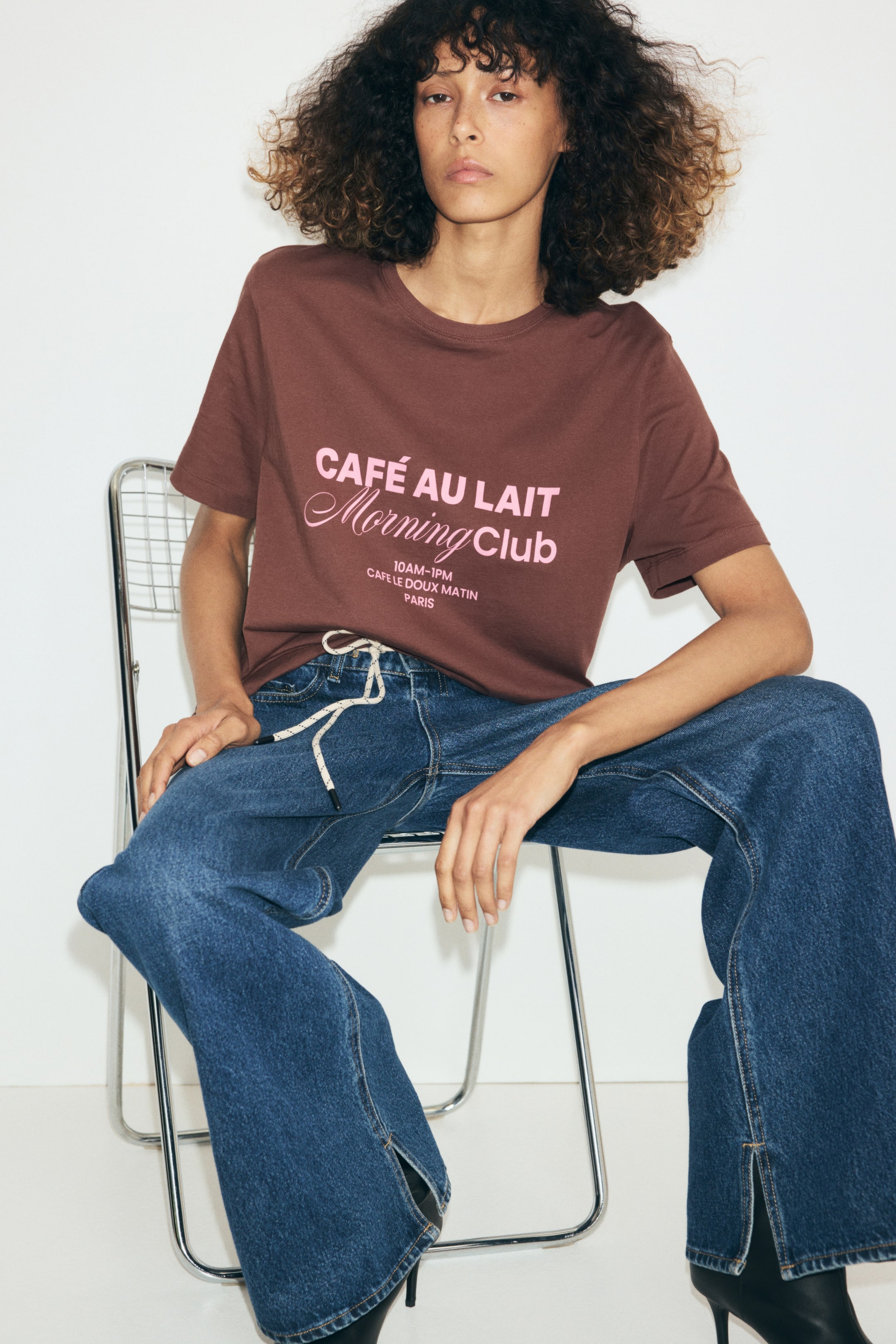 Ampliar la imagen: La modelo lleva puesta una playera de algodón café con cuello redondo, hombros ligeramente caídos y texto gráfico rosa al frente. La playera se combina con jeans azul de pierna ancha con cordón ajustable.